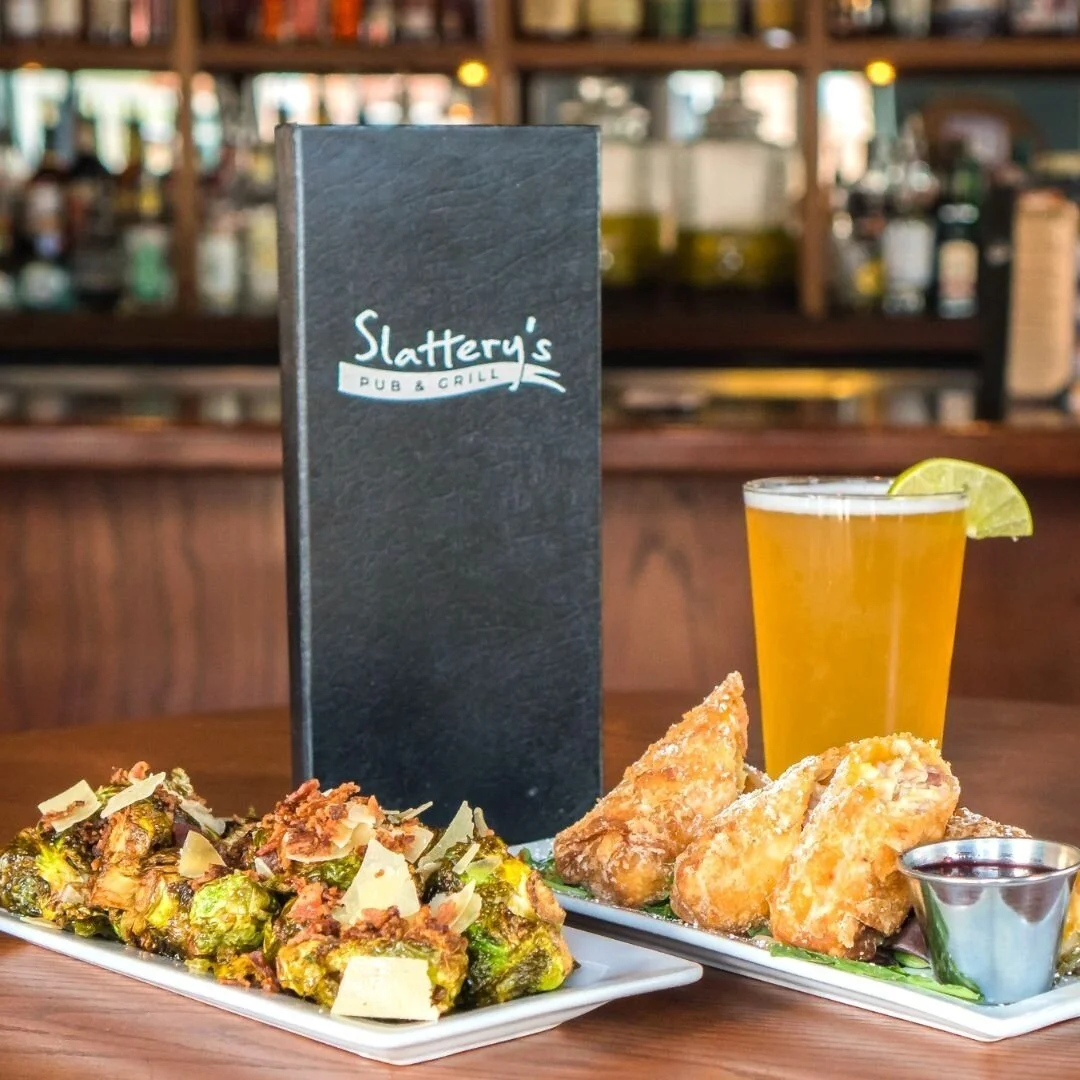 A table at a restaurant called Slattery's Pub & Grill with a menu, a plate of fried chicken wings with dipping sauce, a plate of roasted Brussels sprouts with shaved cheese, and a glass of beer with a lemon wedge.