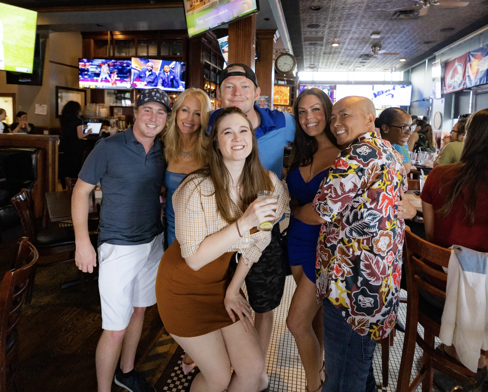 Group of six friends smiling and posing together at a restaurant, with TVs showing sports in the background.