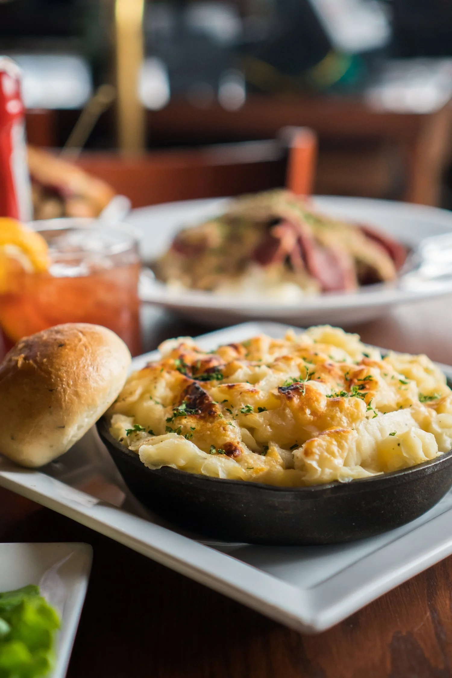 Baked macaroni and cheese in a black skillet with a roll of bread on a white serving platter at a restaurant table.
