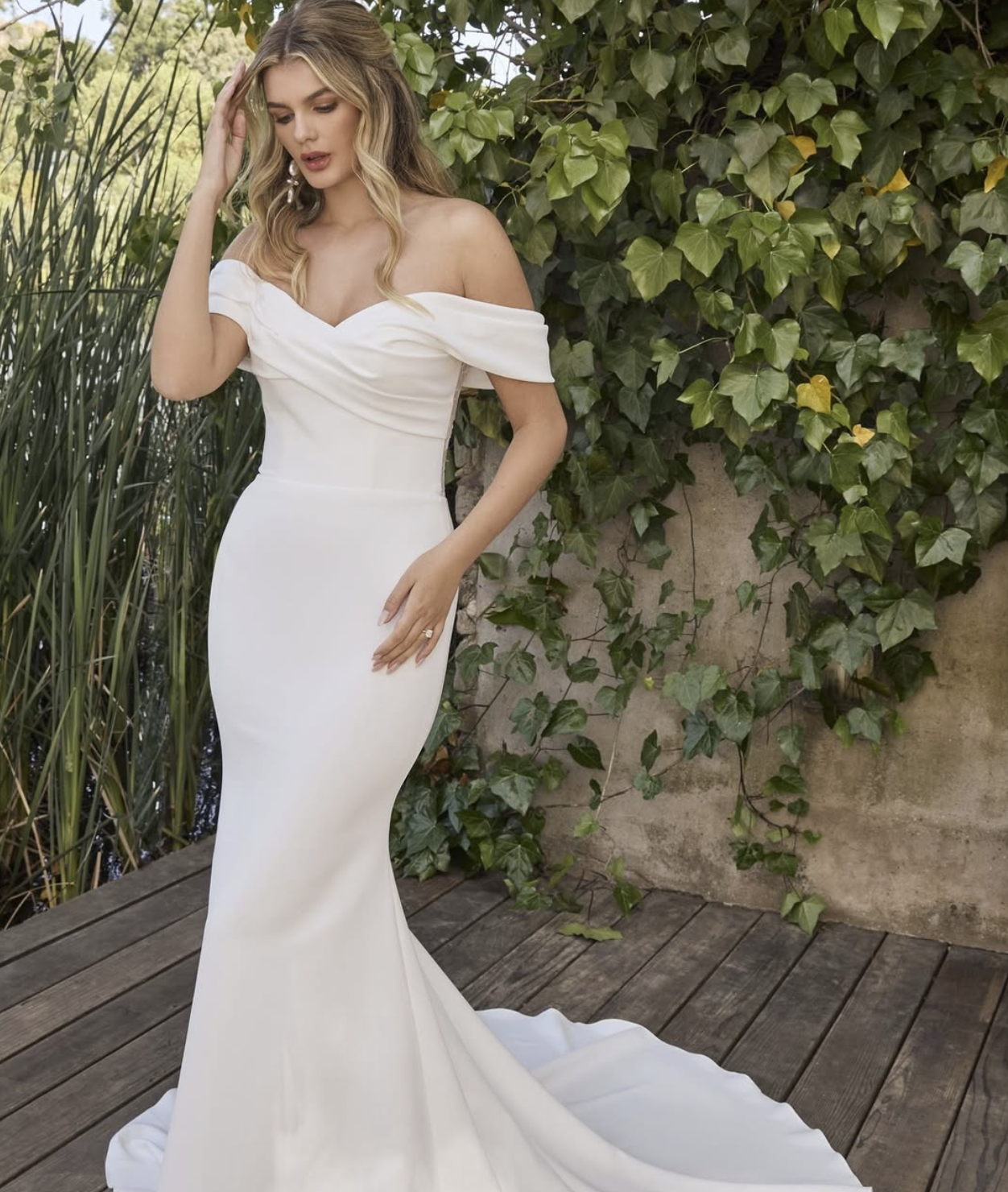 Woman in a white off-shoulder wedding dress standing on a wooden deck surrounded by green foliage.
