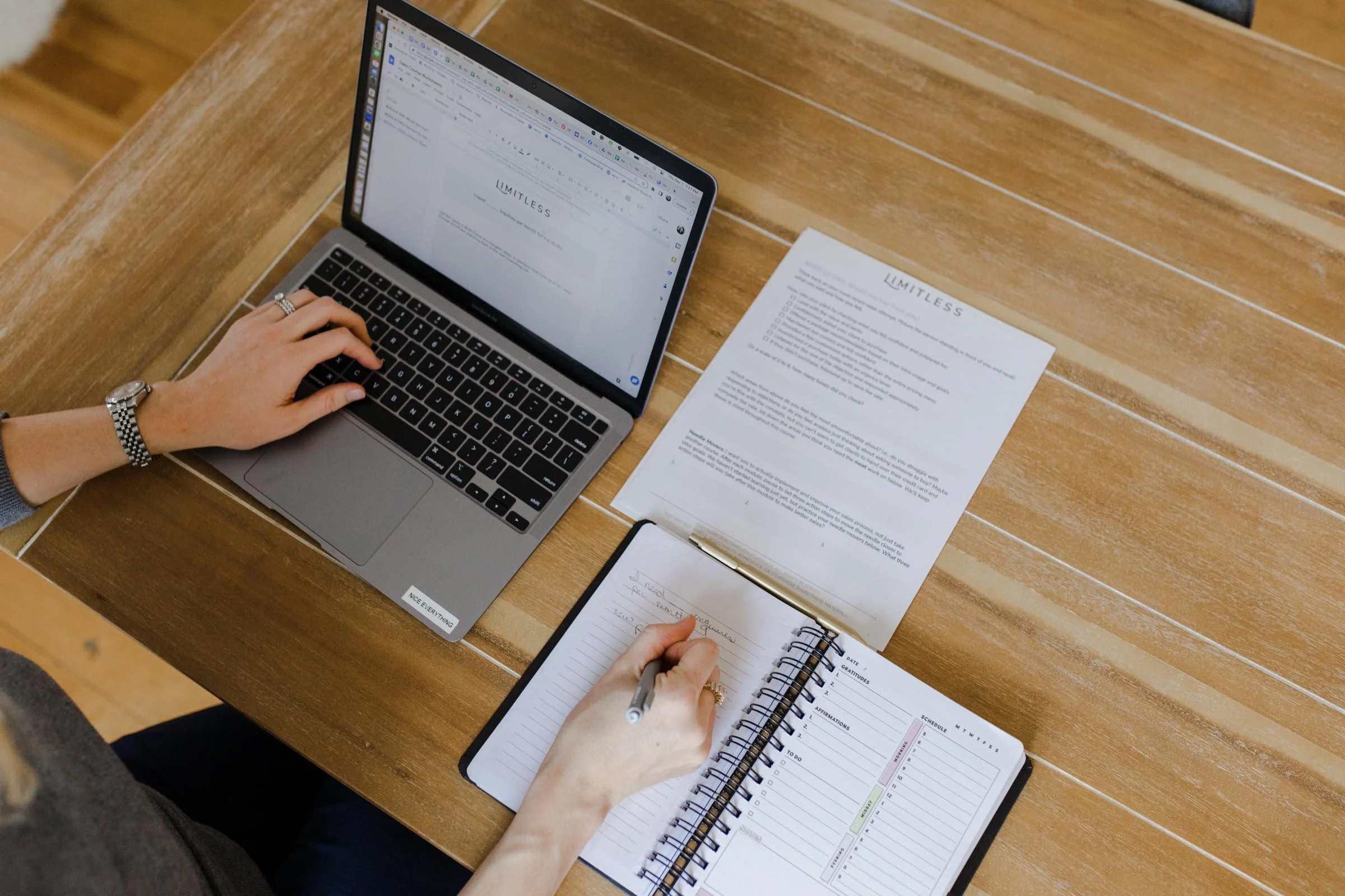 Person working at a wooden desk with a laptop, notebook, and printed document.