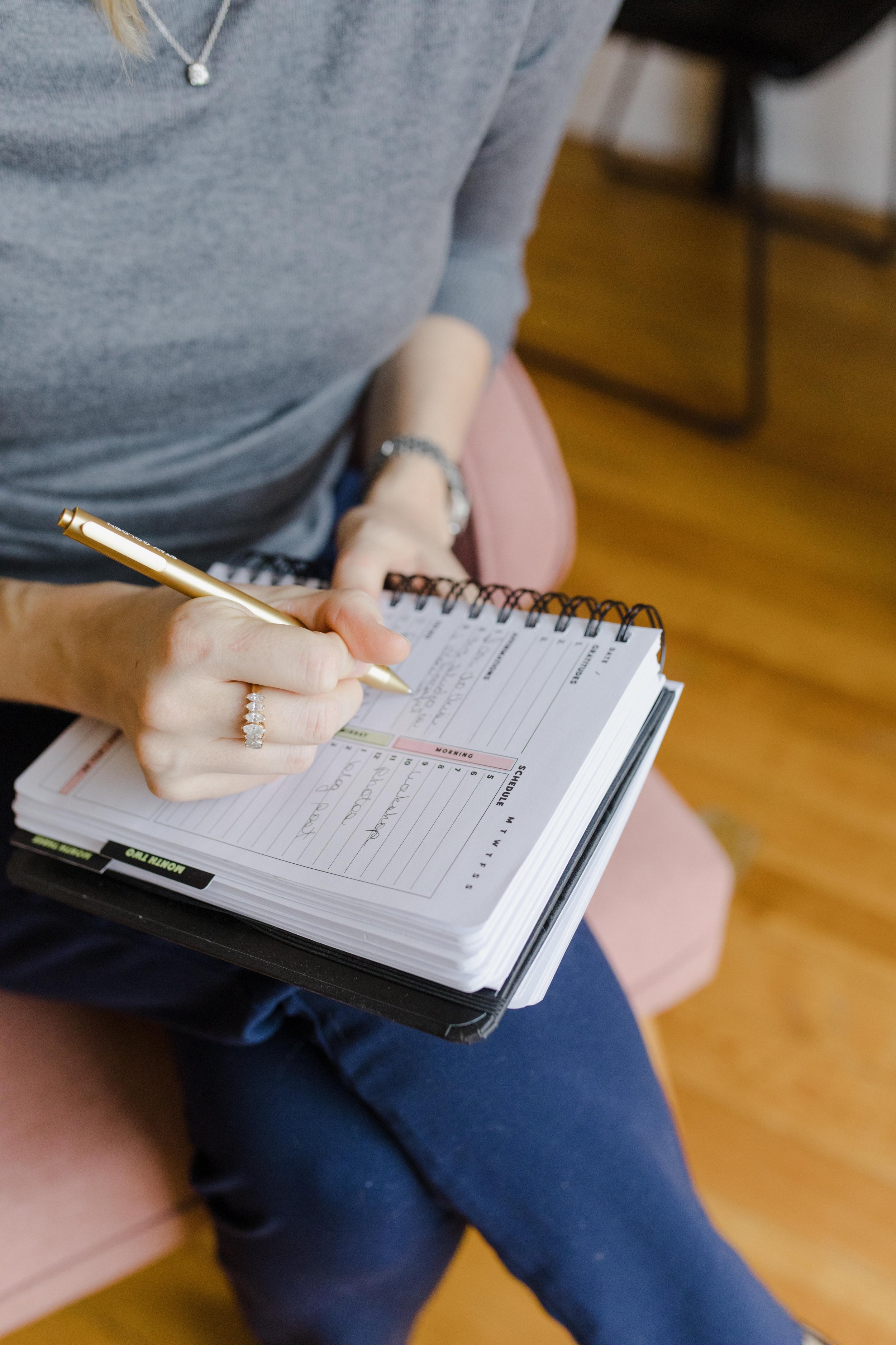 Person writing in a spiral-bound planner with a gold pen, wearing a gray sweater and jeans, sitting on a pink chair.