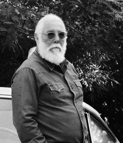 Man with white beard and sunglasses standing outdoors next to a vehicle with dense foliage in the background.