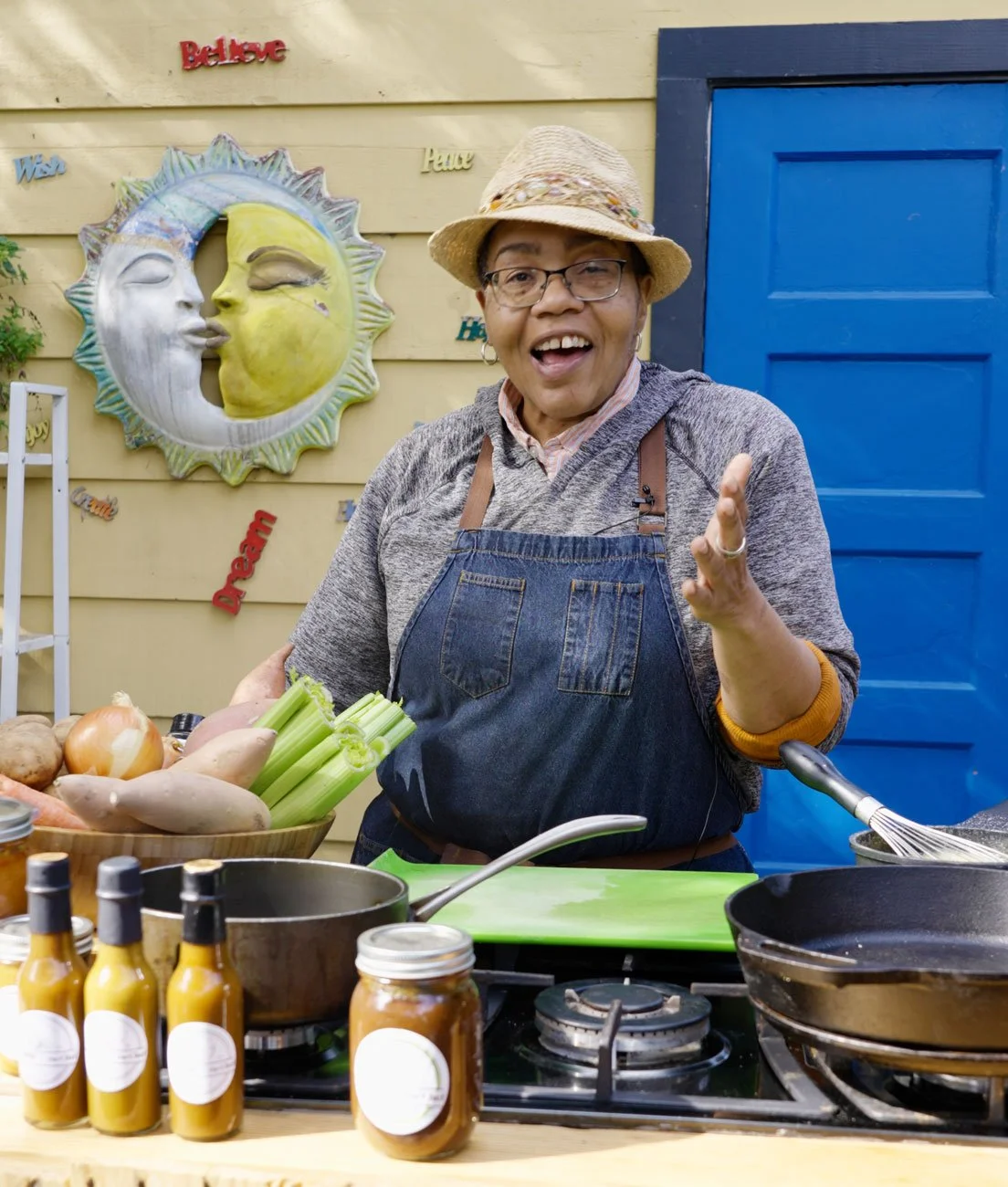 chef Sarah in apron and straw hat set up outside for a cooking demo with a stove top and a bowl of vegetables. standing in front of a blue door and metal art of the sun and the moon kissing