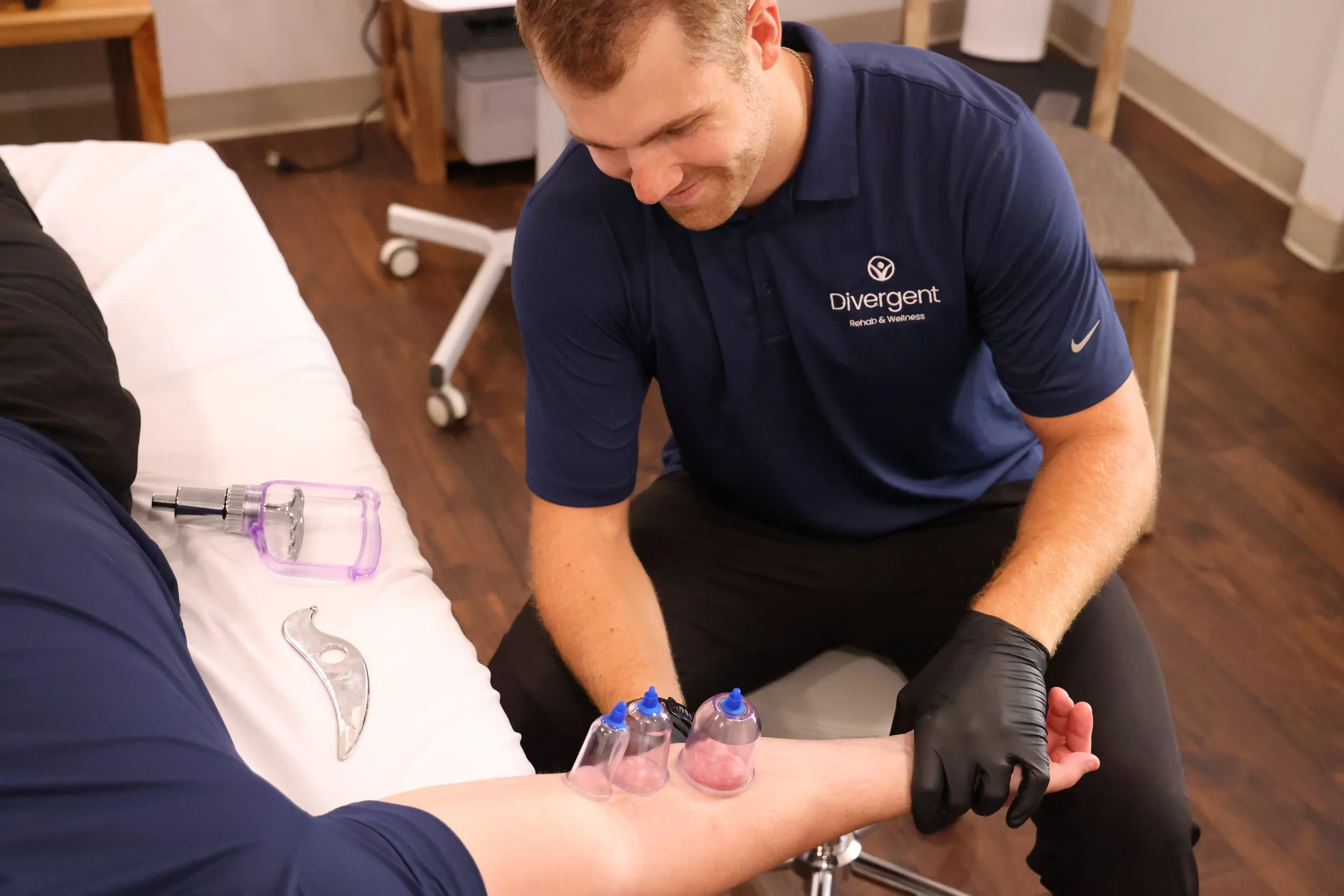A healthcare professional in a navy polo shirt with "Divergent Rehab & Wellness" and a Nike logo on it is providing physical therapy to a patient on a medical table. The patient’s arm is being treated with cupping therapy, and there are syringes with blue caps on the table. The setting appears to be a clinic with wooden flooring and medical equipment in the background.