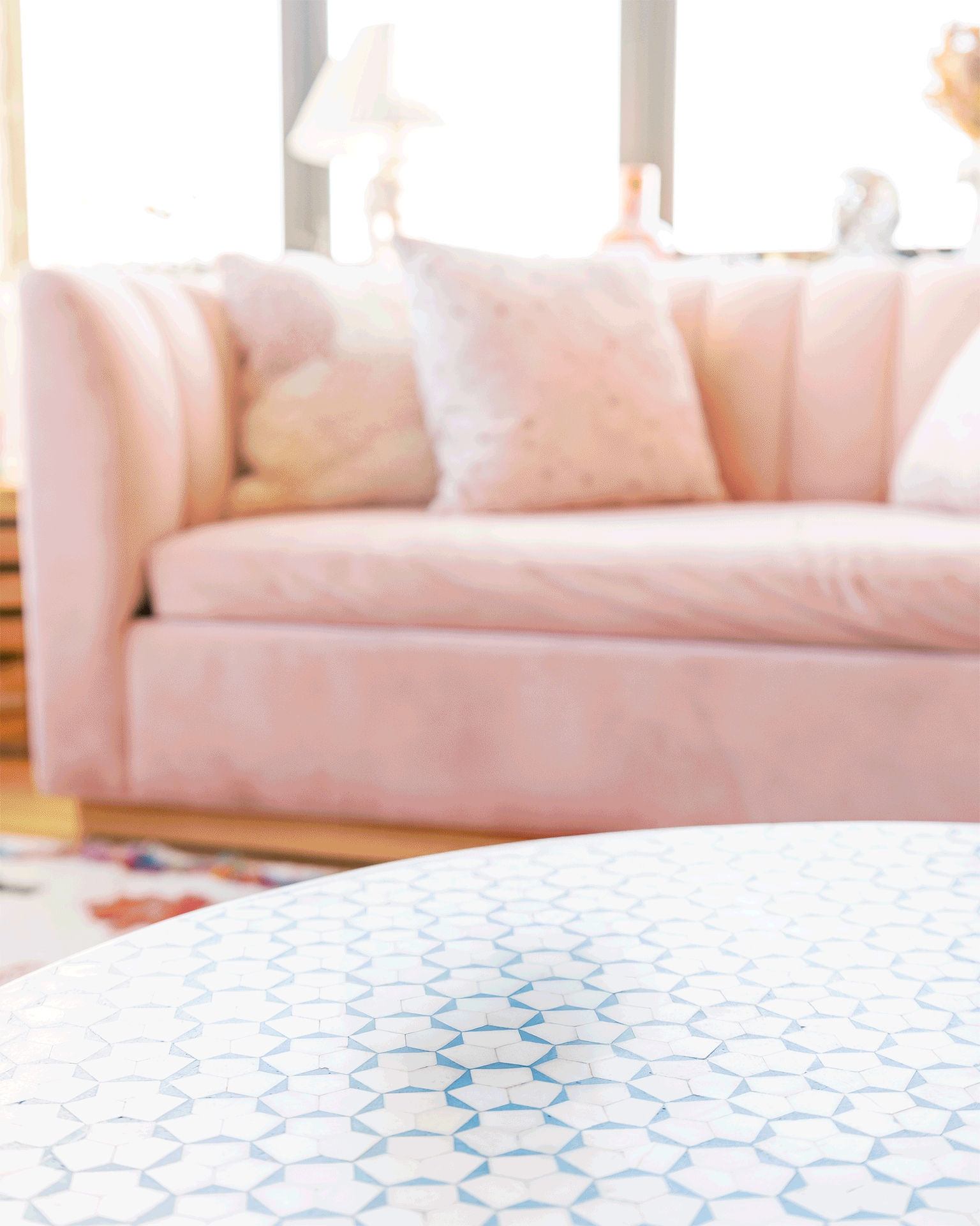 A pastel pink velvet sofa with light-colored pillows in a sunlit living room, with a white and blue patterned table in the foreground. The GIF shows Companion Candles being stacked and unstacked. 