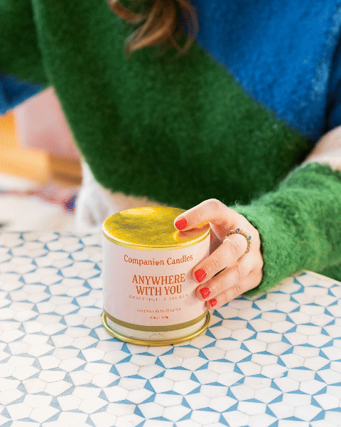 Person with red nail polish holding a candle labeled 'Companion Candles Anywhere With You' on a patterned table.