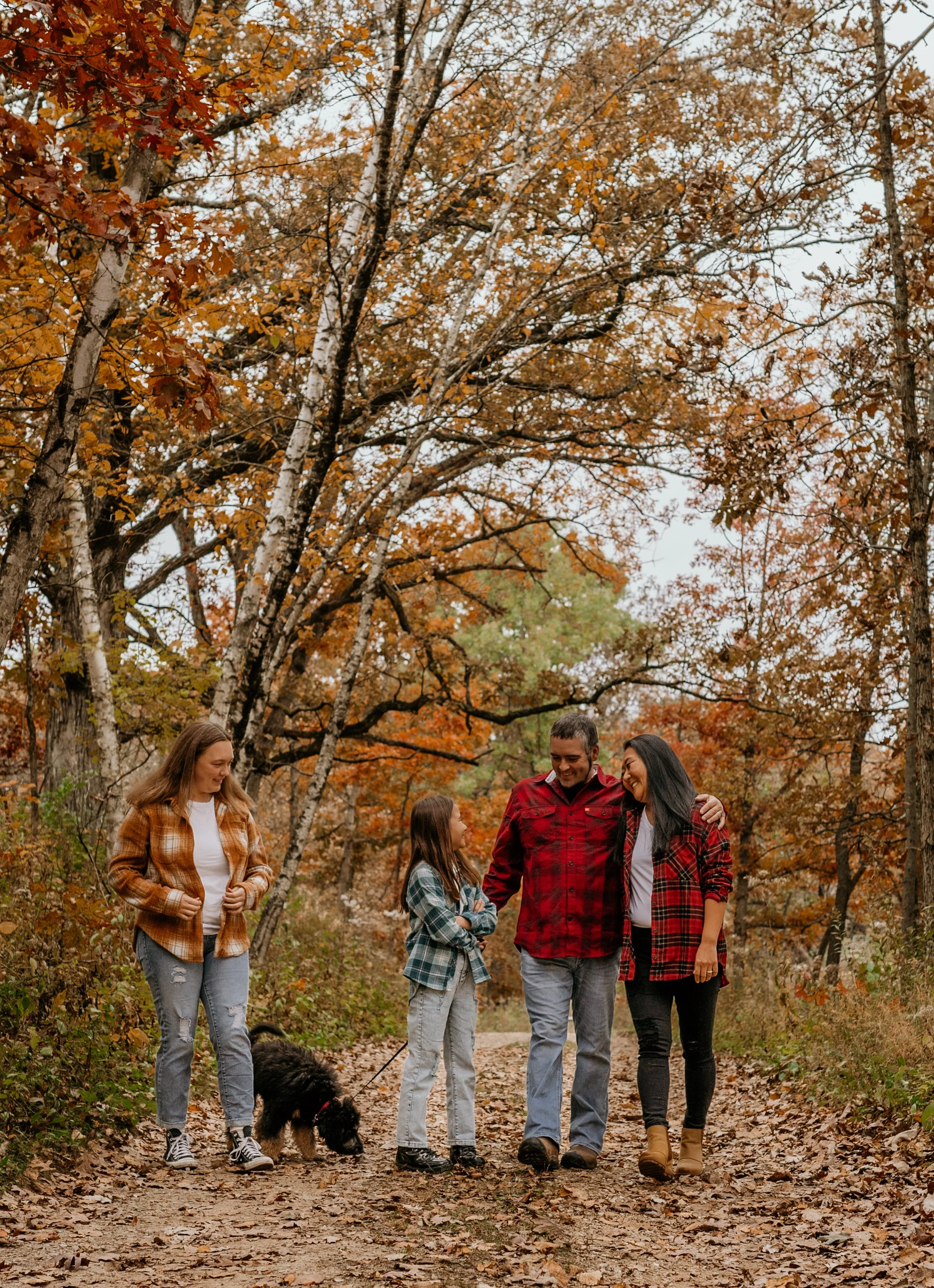 Family of four wearing flannels standing on path with fall trees and their black doodle dog