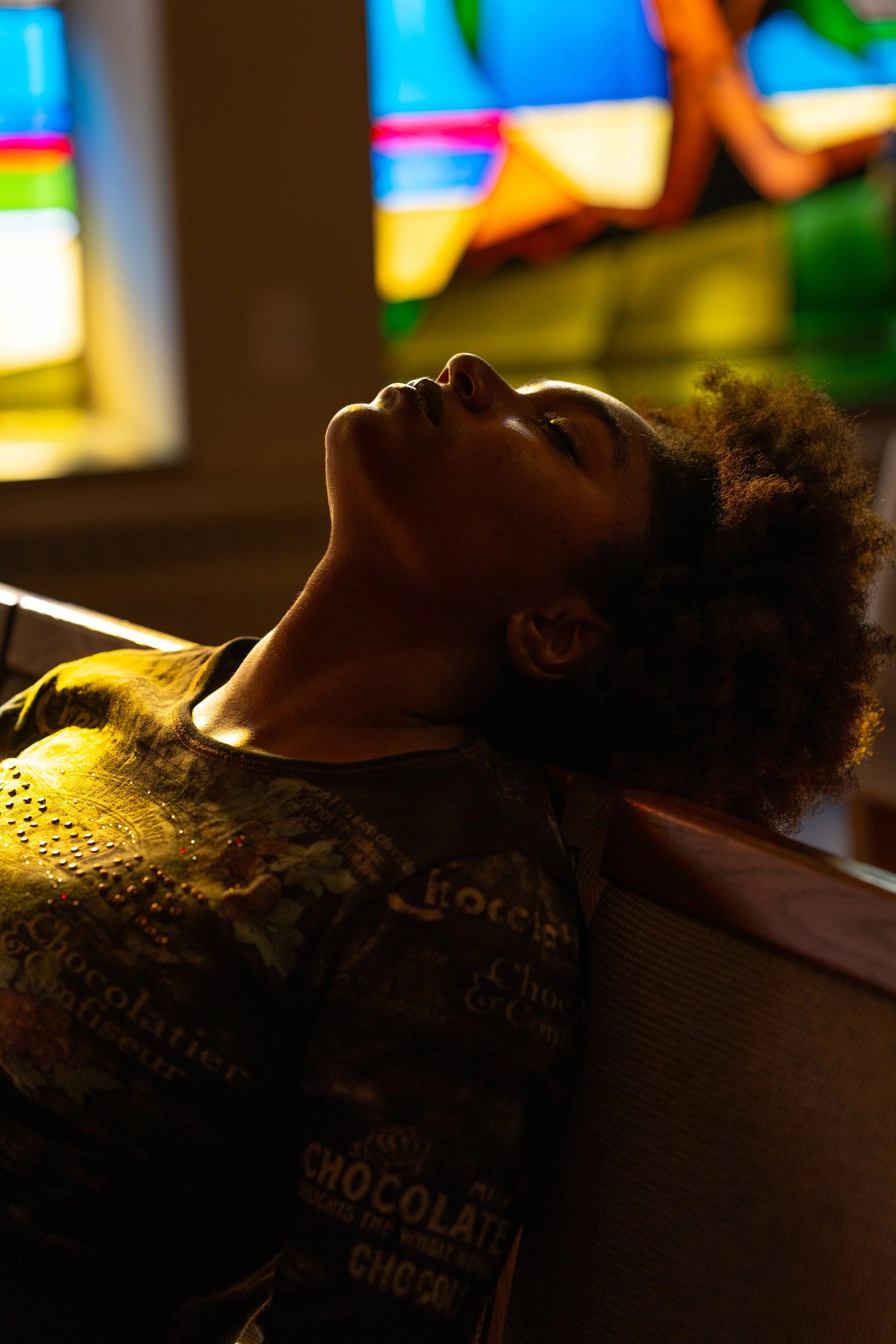 Black woman resting head back on church bench with stain glass window light shinning on her face