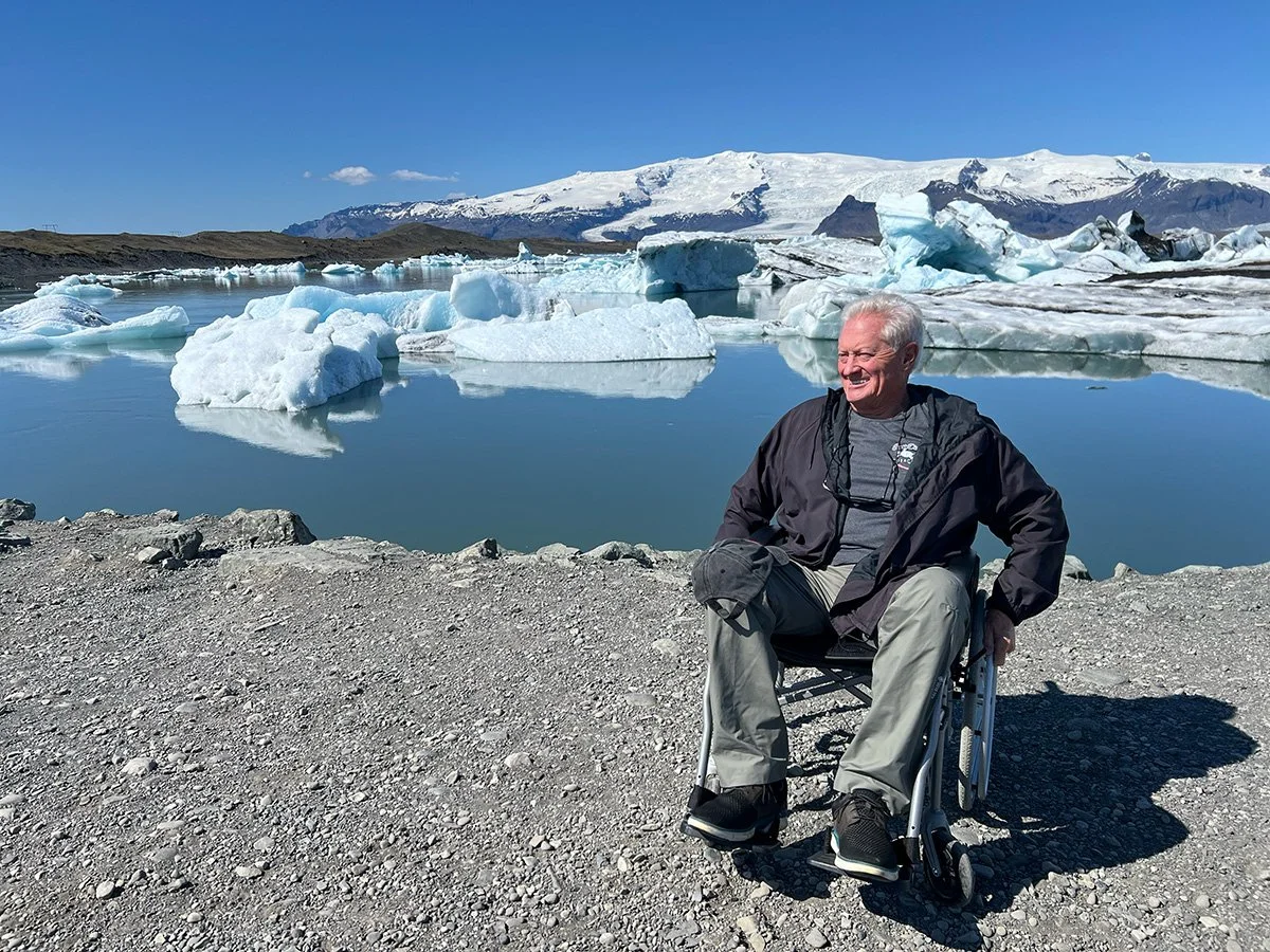 An elderly man in a wheelchair smiling and sitting outdoors near a lake with icebergs, snow-capped mountains, and a blue sky in the background.