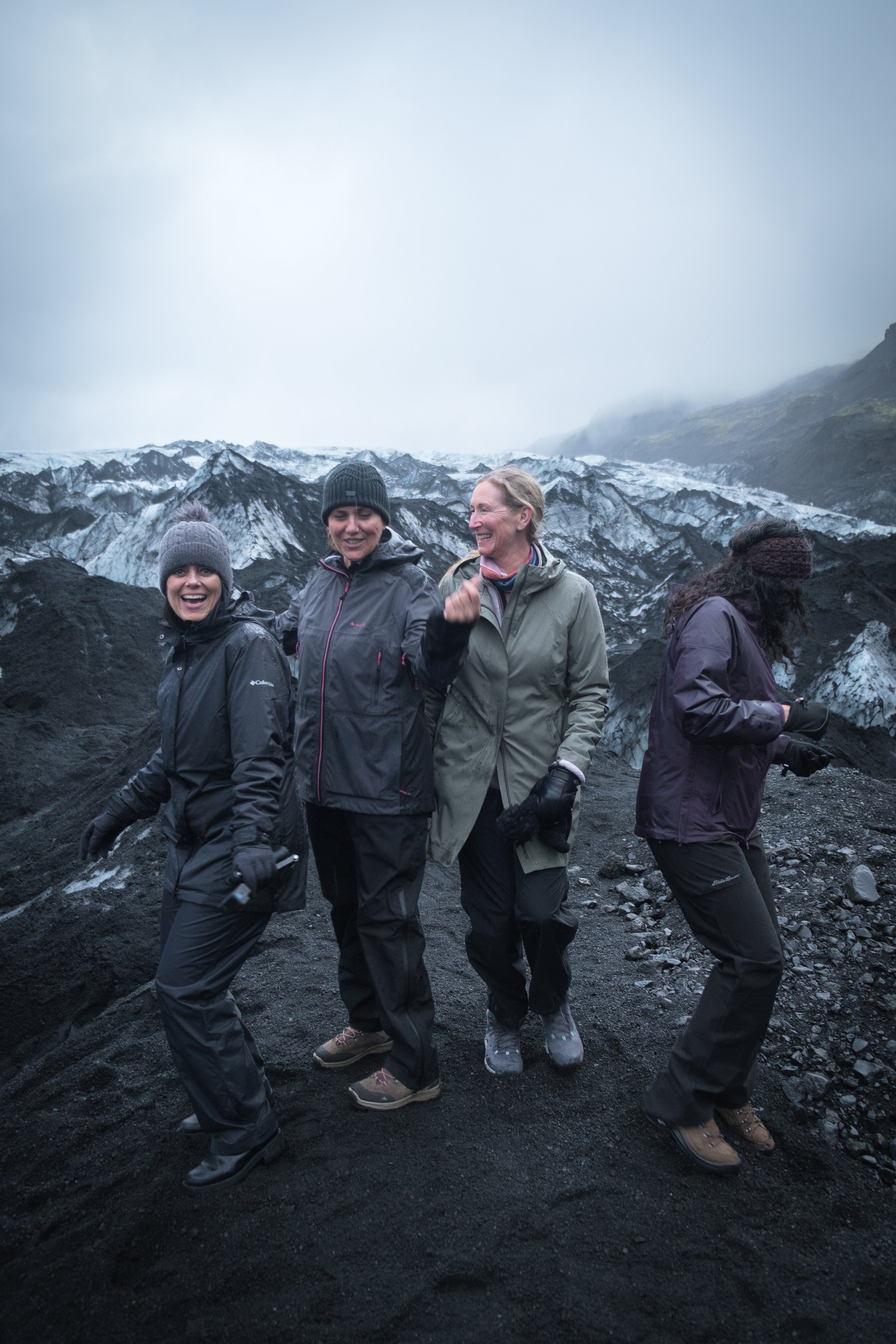 Four women standing on volcanic rocks, dressed in outdoor gear, smiling and interacting with each other, with a snow-covered glacier and mountains in the background.
