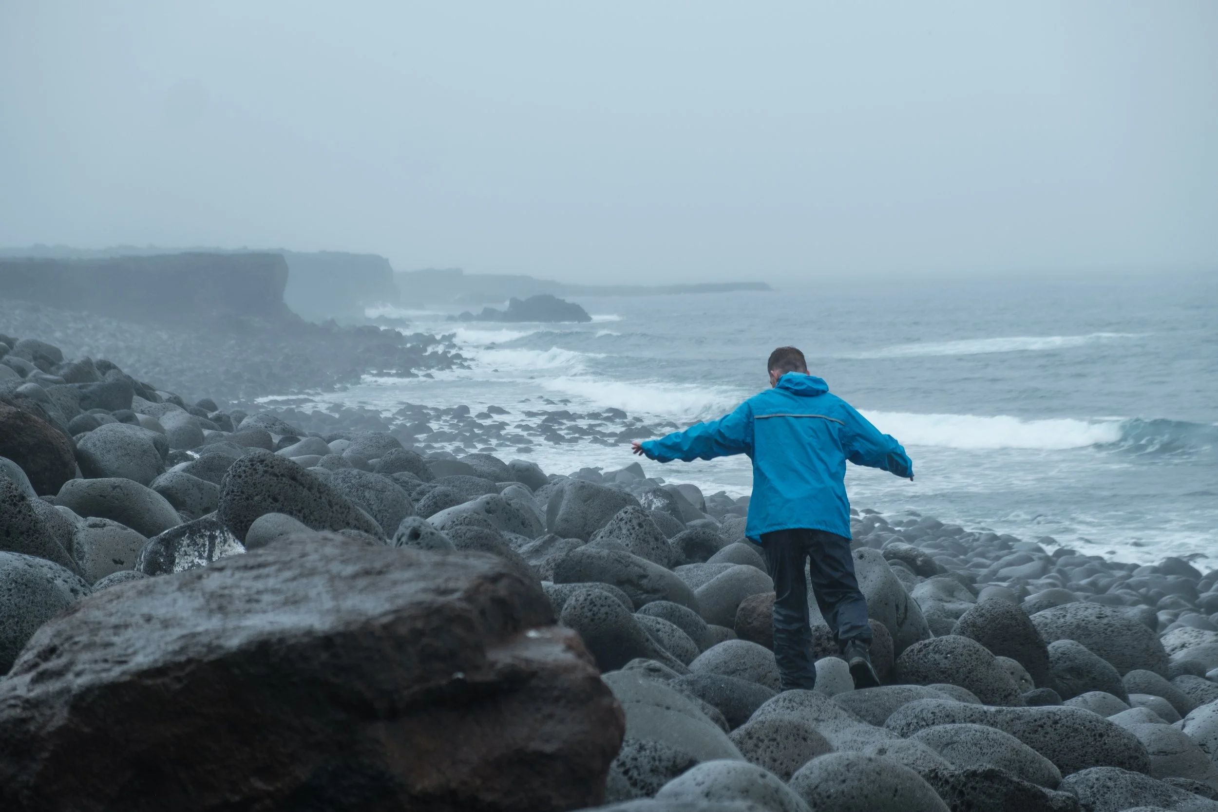 A person wearing a blue jacket and black pants walking on a rocky beach near the ocean with waves and cliffs in the background on a foggy day.