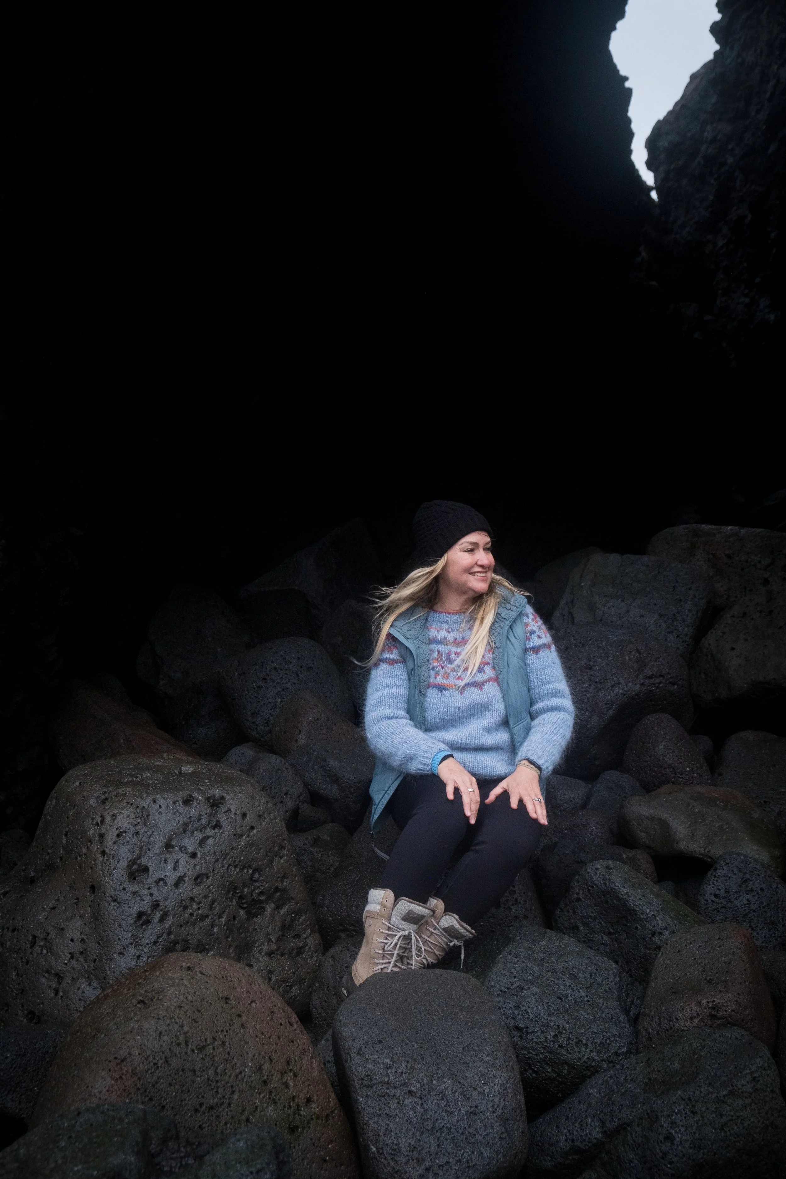 A woman with long blonde hair, dressed in a colorful sweater, gray vest, and beige hiking boots, smiling and sitting among large volcanic rocks in a dark cave or crevice with an opening to the sky visible in the background.