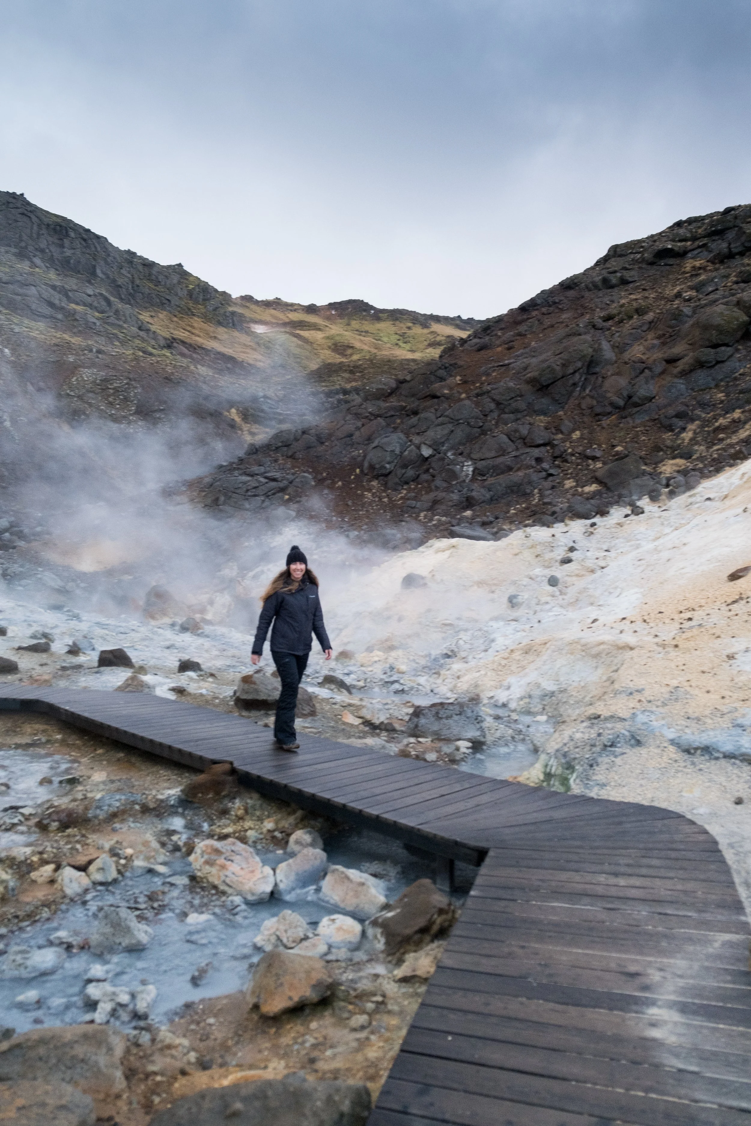 A woman walking on a wooden trail over rocky, steaming terrain with mountains in the background and a cloudy sky above.