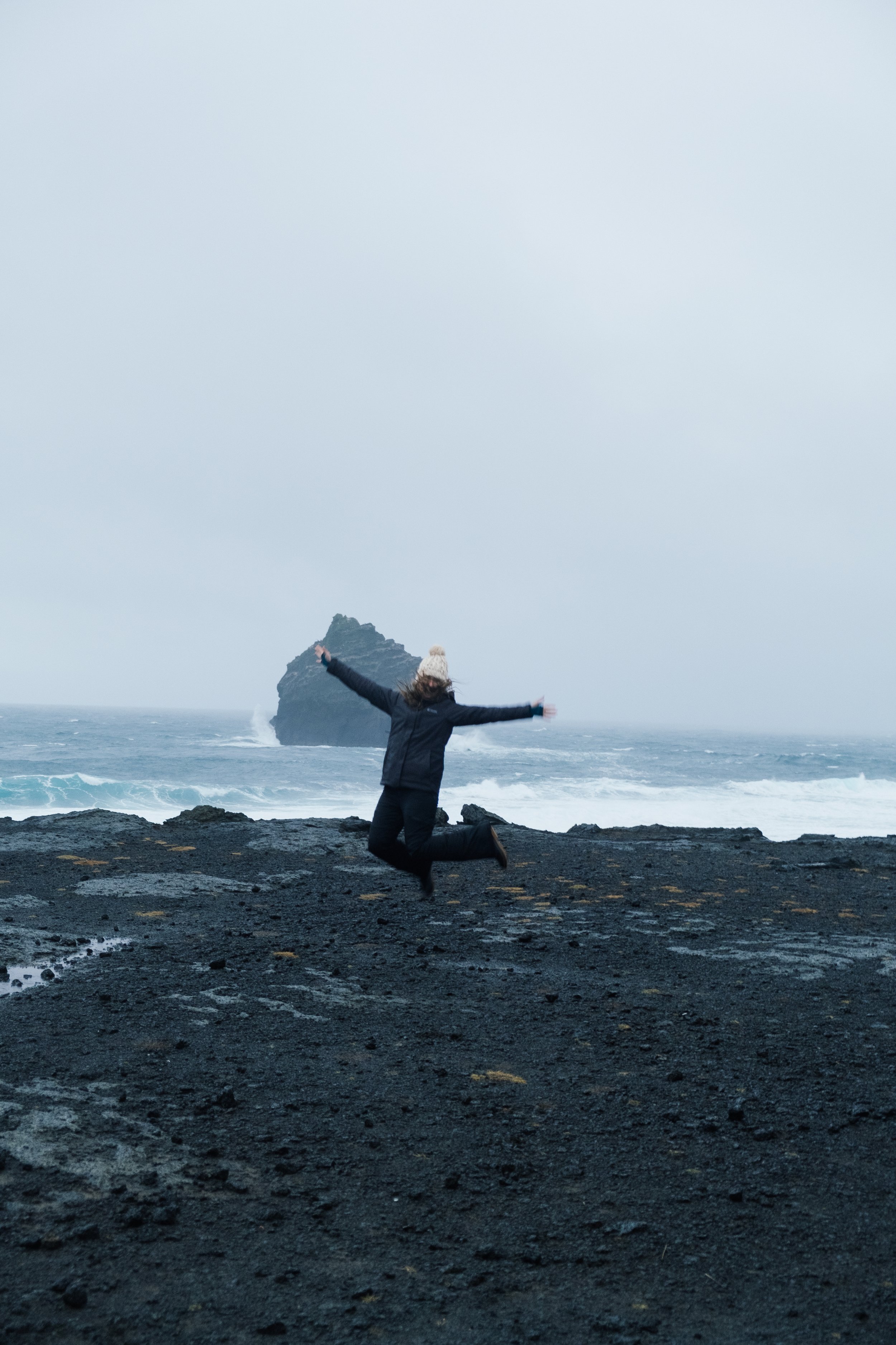 A person kneeling on a rocky black beach with arms outstretched, facing the ocean with waves crashing and a large rock formation in the distance under a cloudy sky.