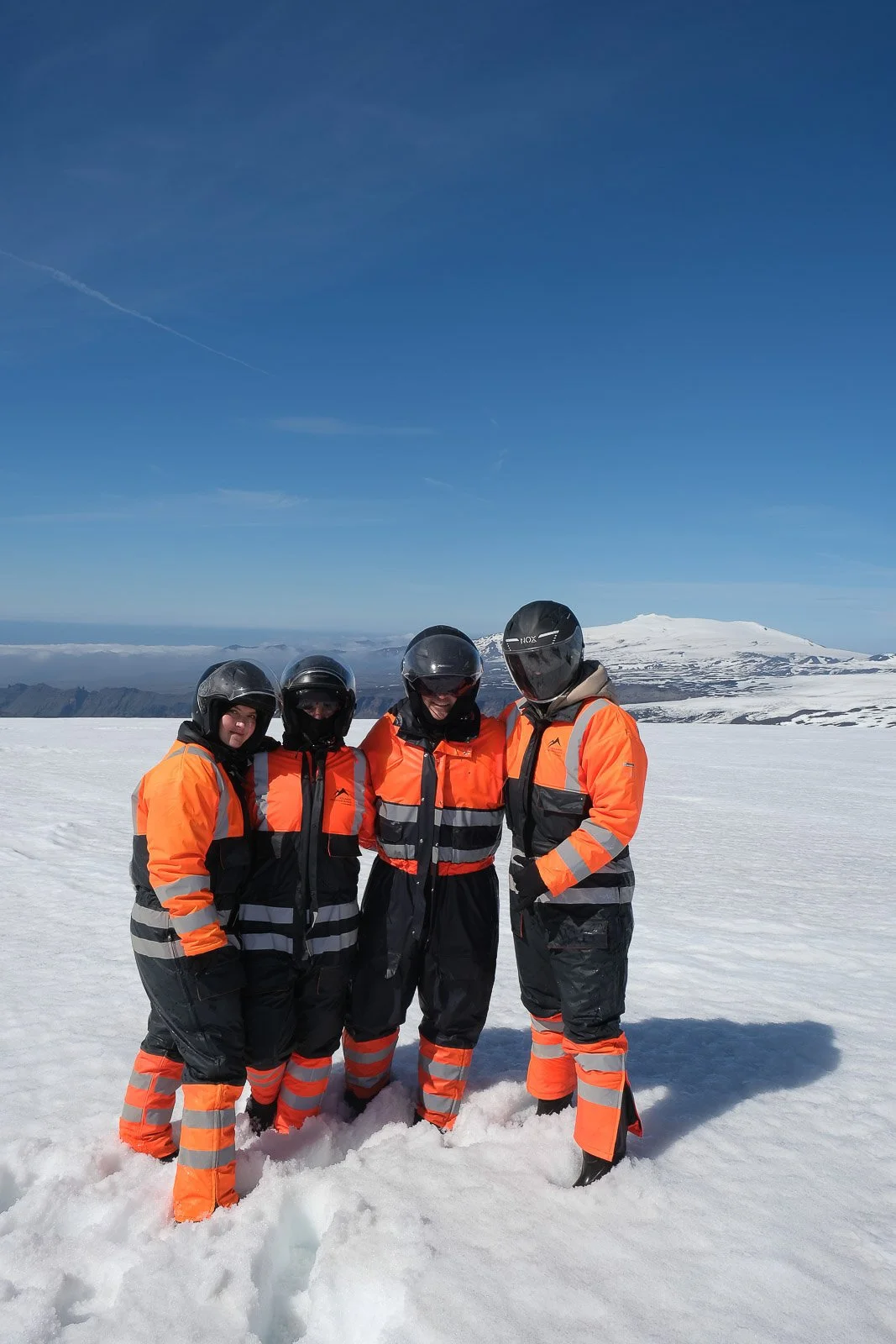 Four people in orange and black cold-weather suits and helmets standing together on snowy terrain, with a snow-covered mountain in the background under a clear blue sky.