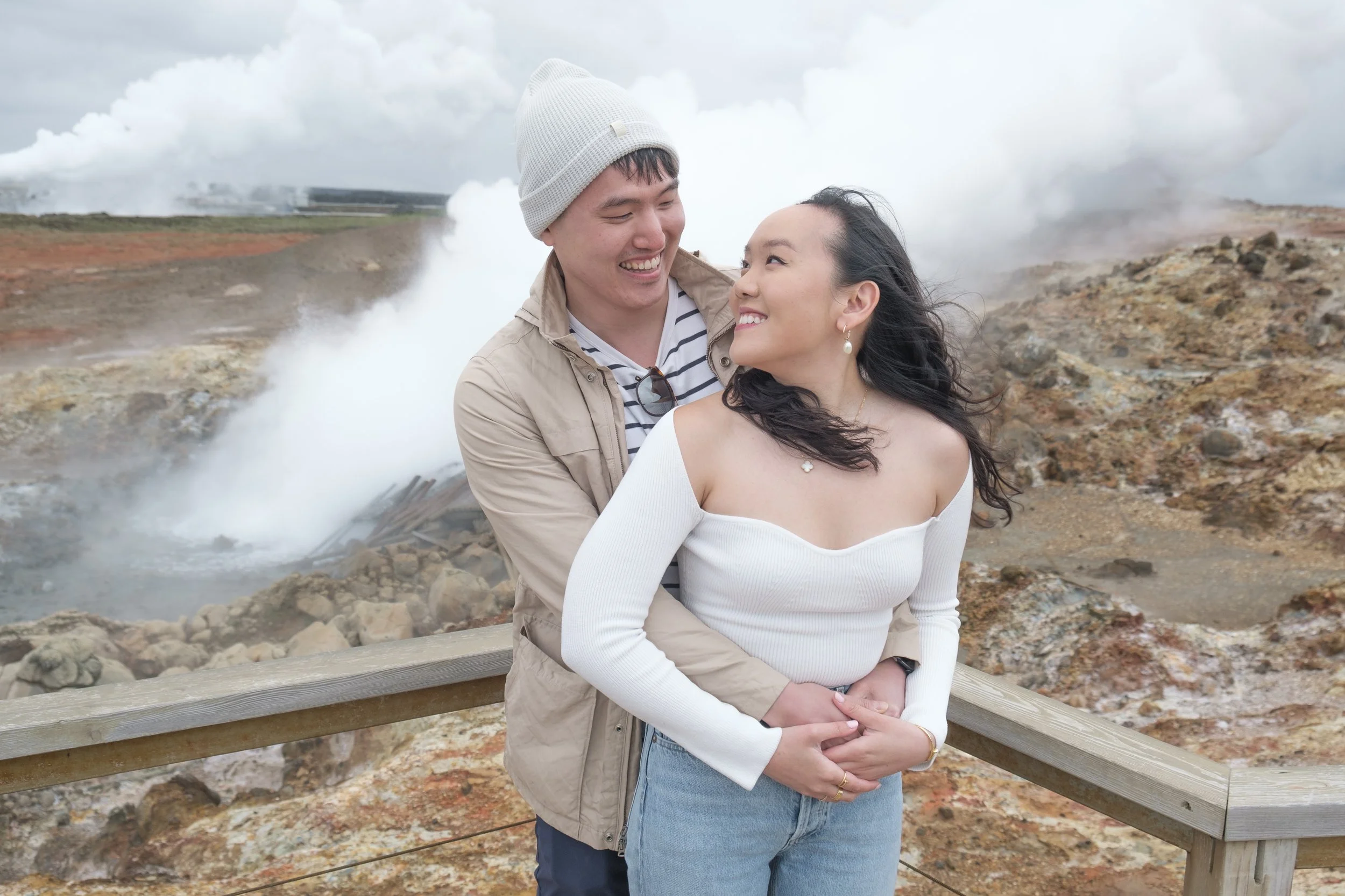A smiling couple standing outdoors near a wooden railing with steam vents in the background, suggesting a geothermal area.