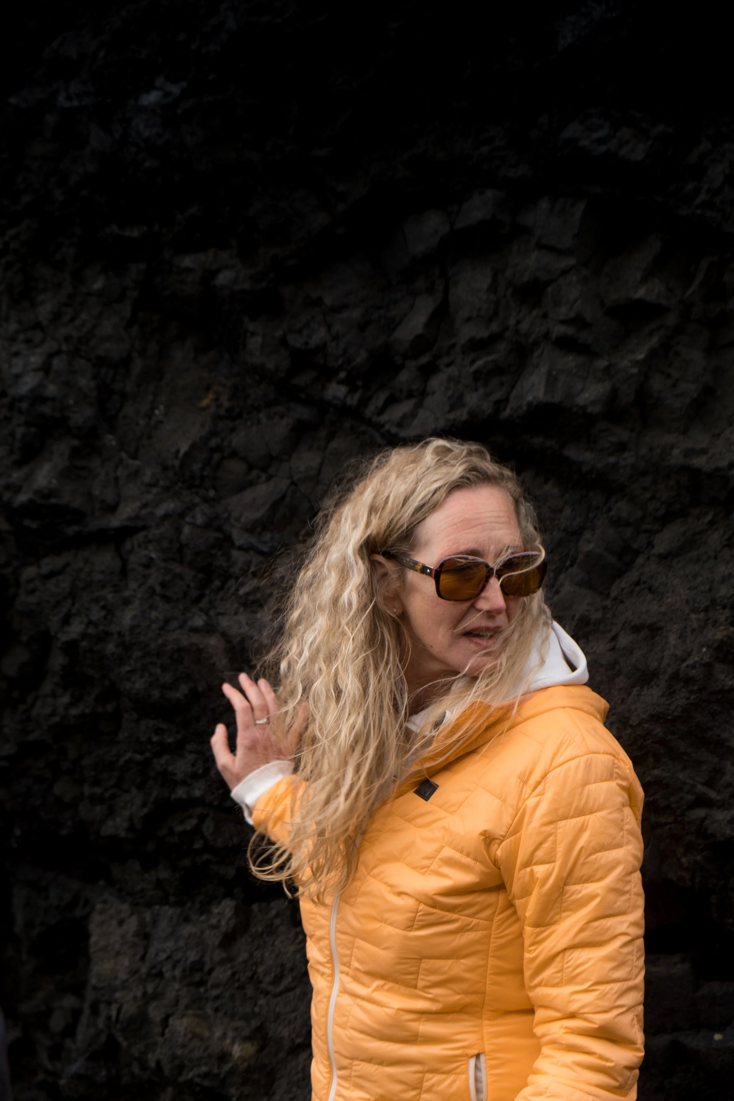 A woman with curly blonde hair wearing sunglasses and an orange jacket, standing outdoors in front of a dark rocky surface.