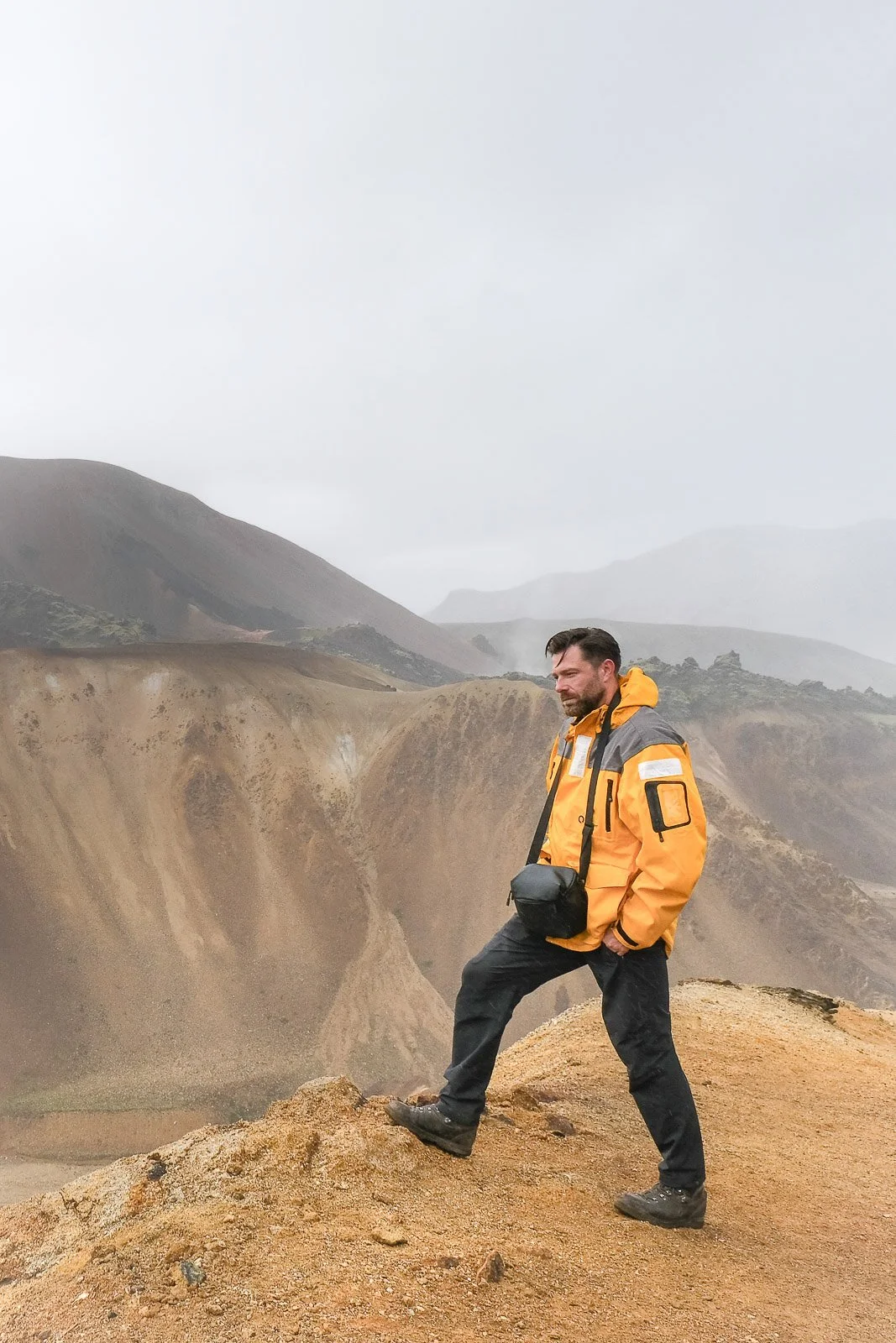 A man in a yellow and gray jacket stands on rocky terrain with mountainous landscape in the background, under a cloudy sky.