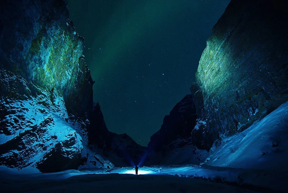 A person standing in a snowy canyon at night illuminated by a flashlight, with tall rock walls on either side and the starry sky overhead.