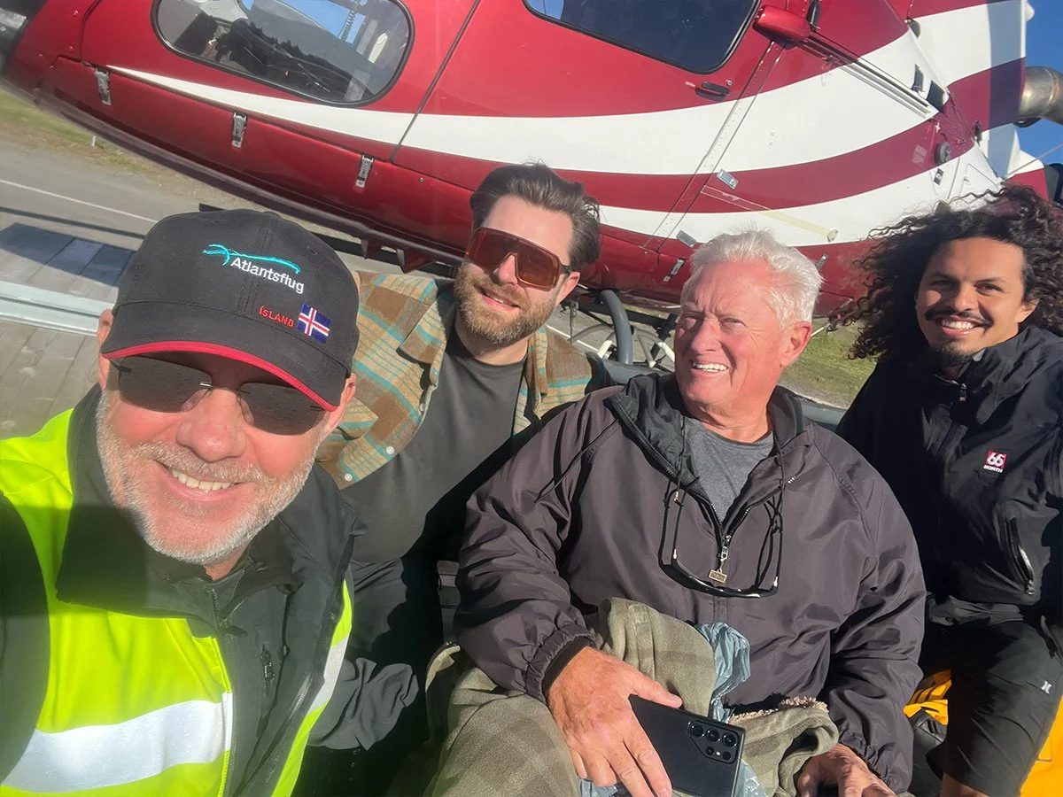 Four men taking a selfie in front of a red helicopter. The man on the far left is wearing a black cap with 'Atlantsflug' and Norwegian flag, and a black and yellow jacket. The man second from the left has dark hair, sunglasses, and a plaid shirt. The third man has white hair, is dressed in a gray jacket, and holding a phone. The man on the far right has curly hair and a black jacket. The background includes part of a road and blue sky.