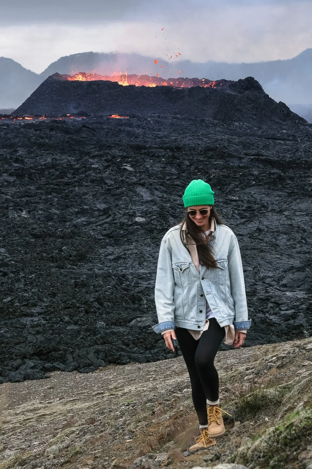 A smiling woman in a green beanie, sunglasses, denim jacket, black leggings, and tan boots hiking against a volcanic landscape with an erupting volcano in the background.