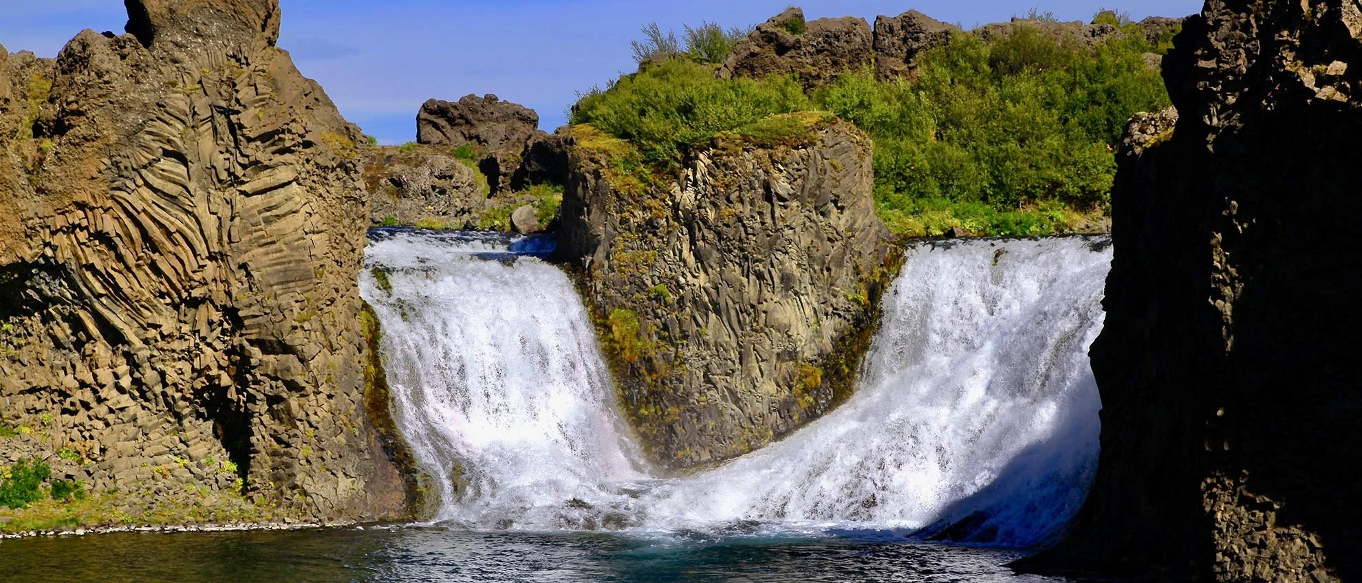 A river flowing over a small waterfall surrounded by rocky cliffs and green vegetation in Hjálparfoss Iceleand