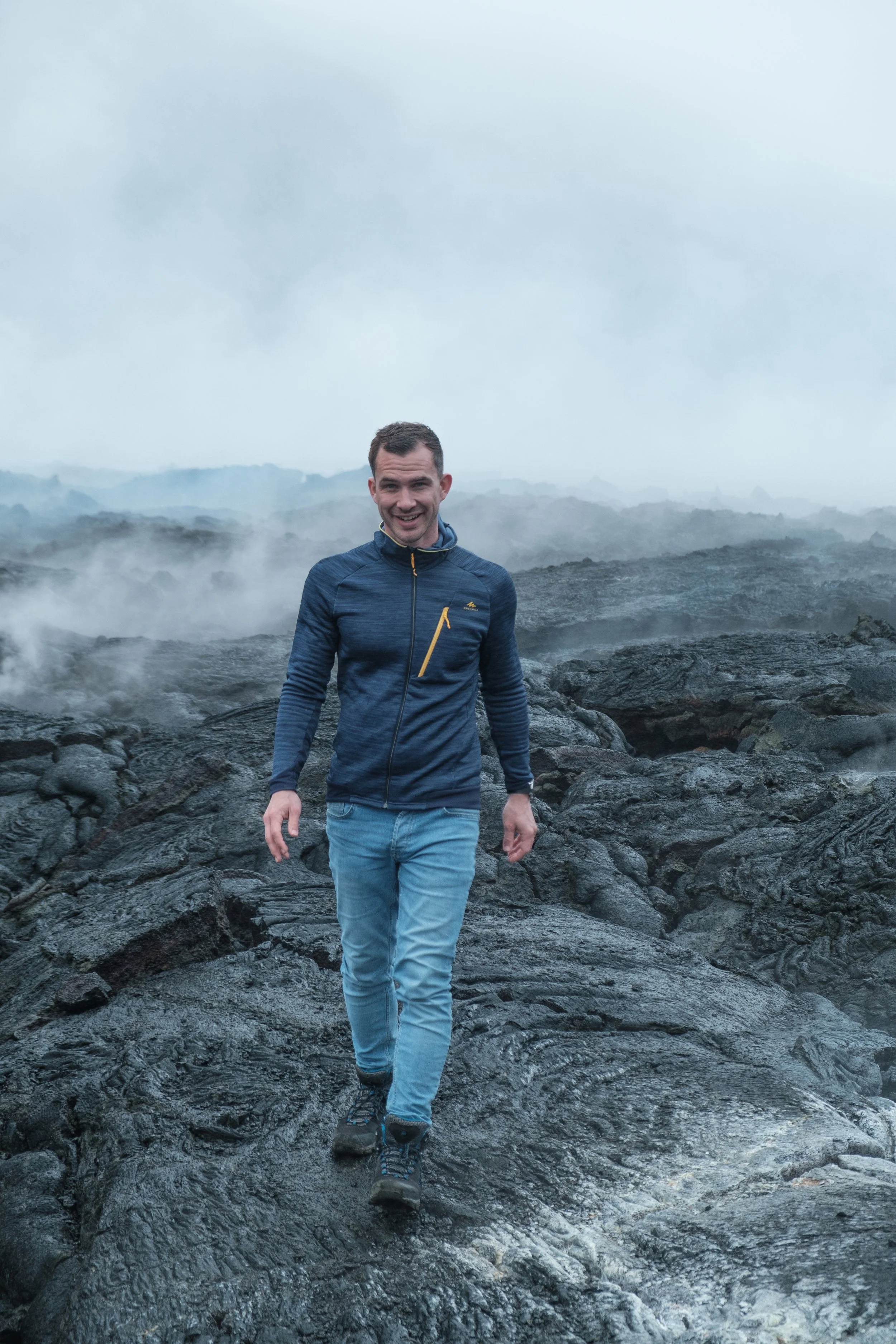 A man in a blue jacket and jeans walking on volcanic rocks with steam rising around him, and a cloudy sky in the background.