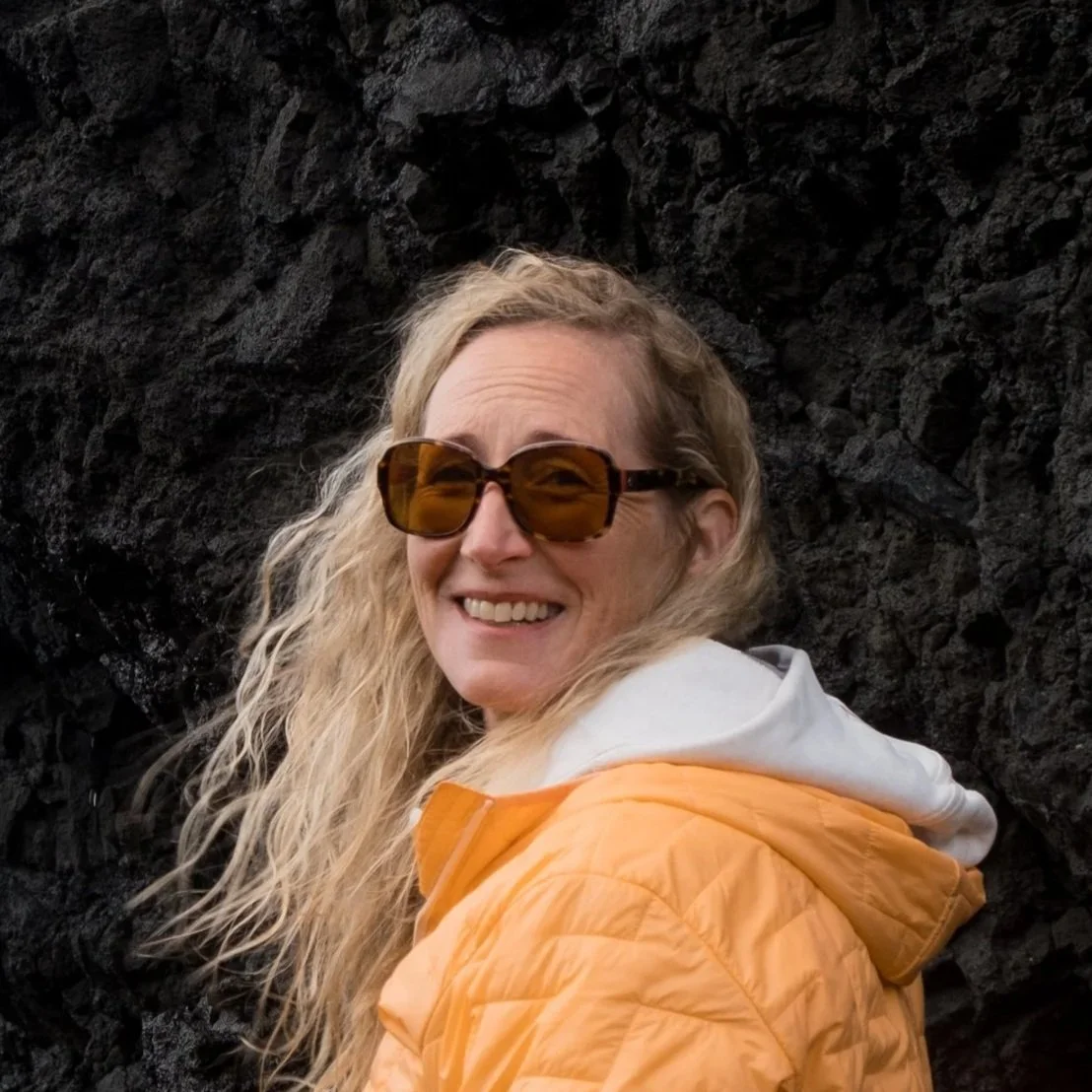 Woman with curly blonde hair wearing sunglasses and a yellow jacket, smiling in front of a dark rocky background.