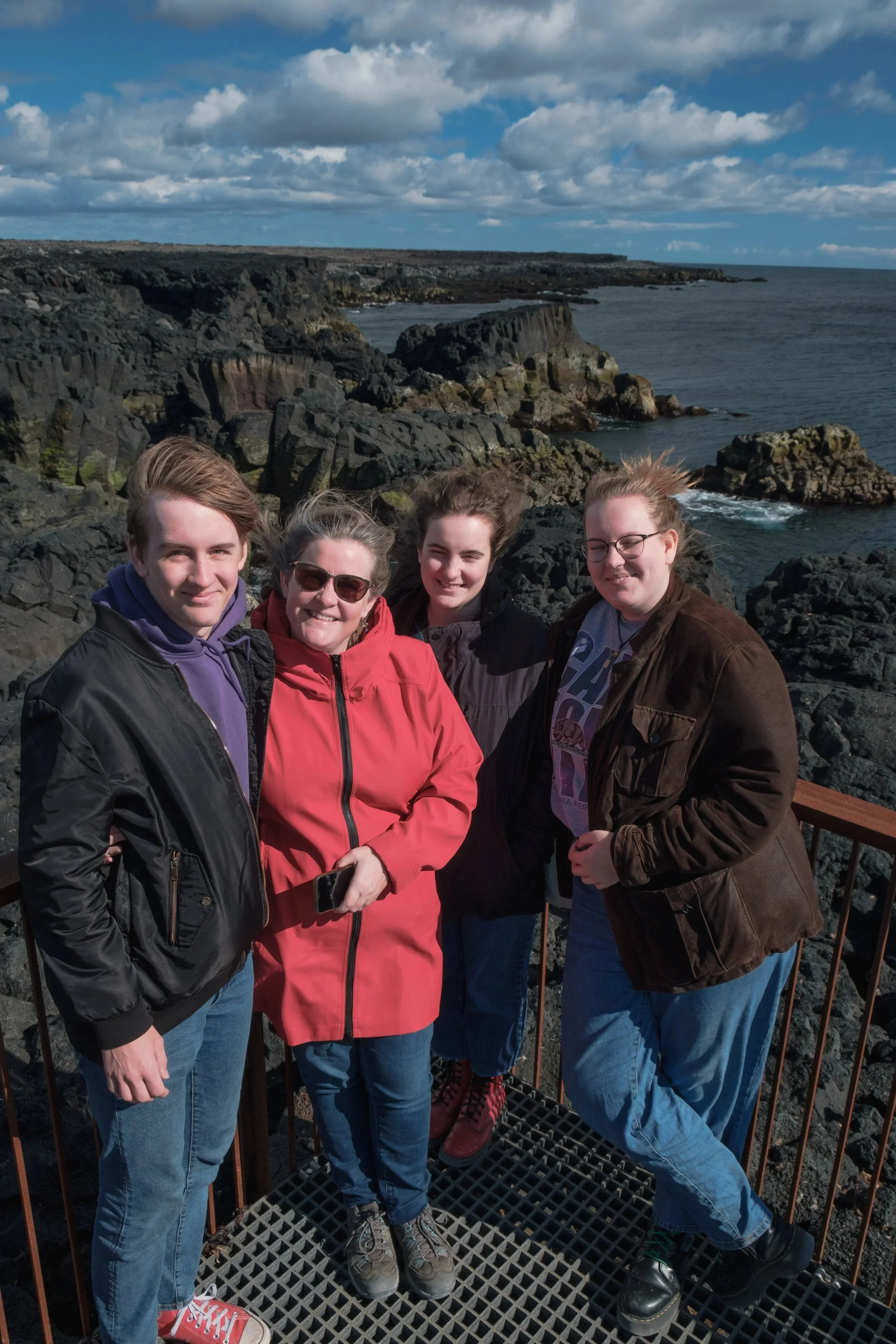 Four people standing on a rocky coast with cliffs and ocean in the background, under a partly cloudy sky, smiling for a photo.