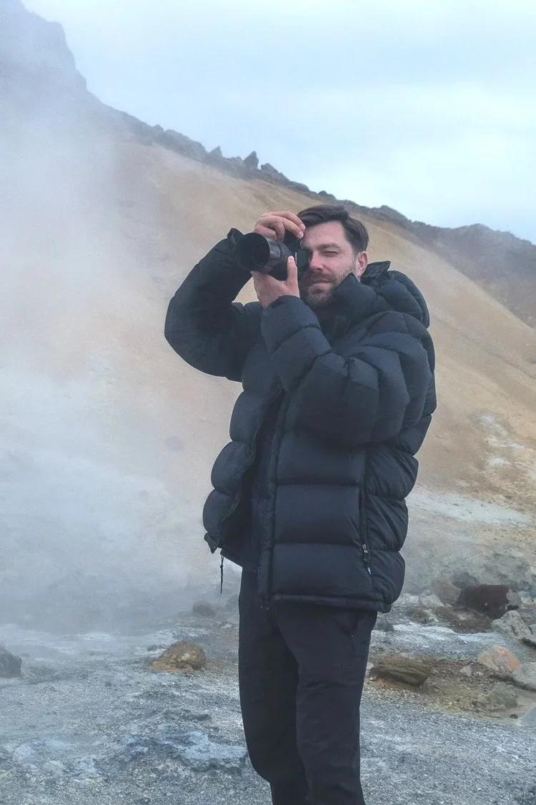 A man in a black puffer jacket taking a photo with a camera in a rocky geothermal area, with steam rising in the background and a hillside in the distance.