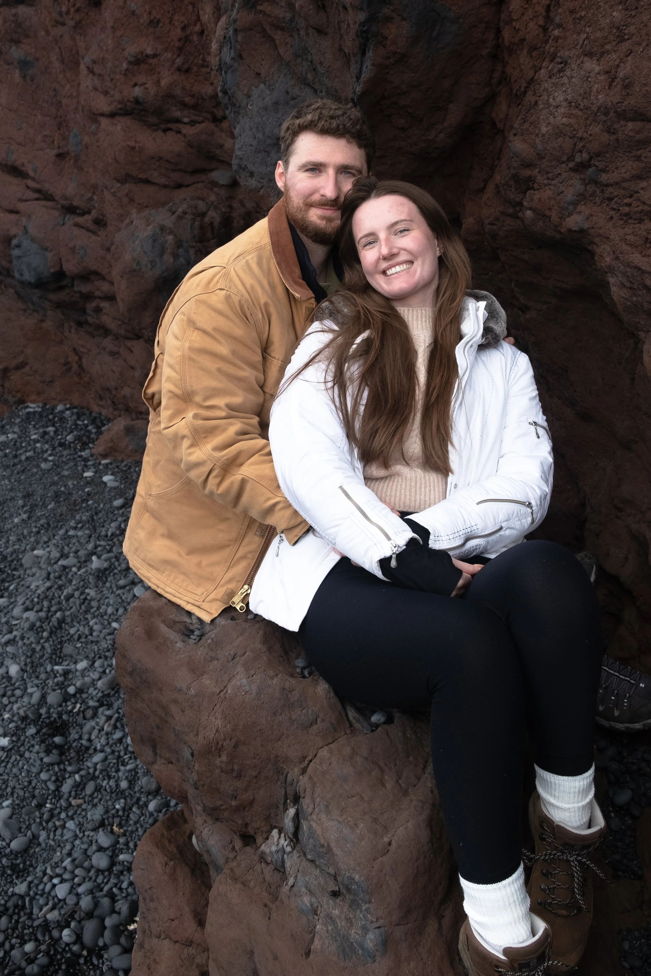 A man and woman sitting on rocks by the ocean. The man has brown hair and a beard, wearing a tan jacket. The woman has long brown hair, wearing a white jacket and black pants. They are smiling at the camera.