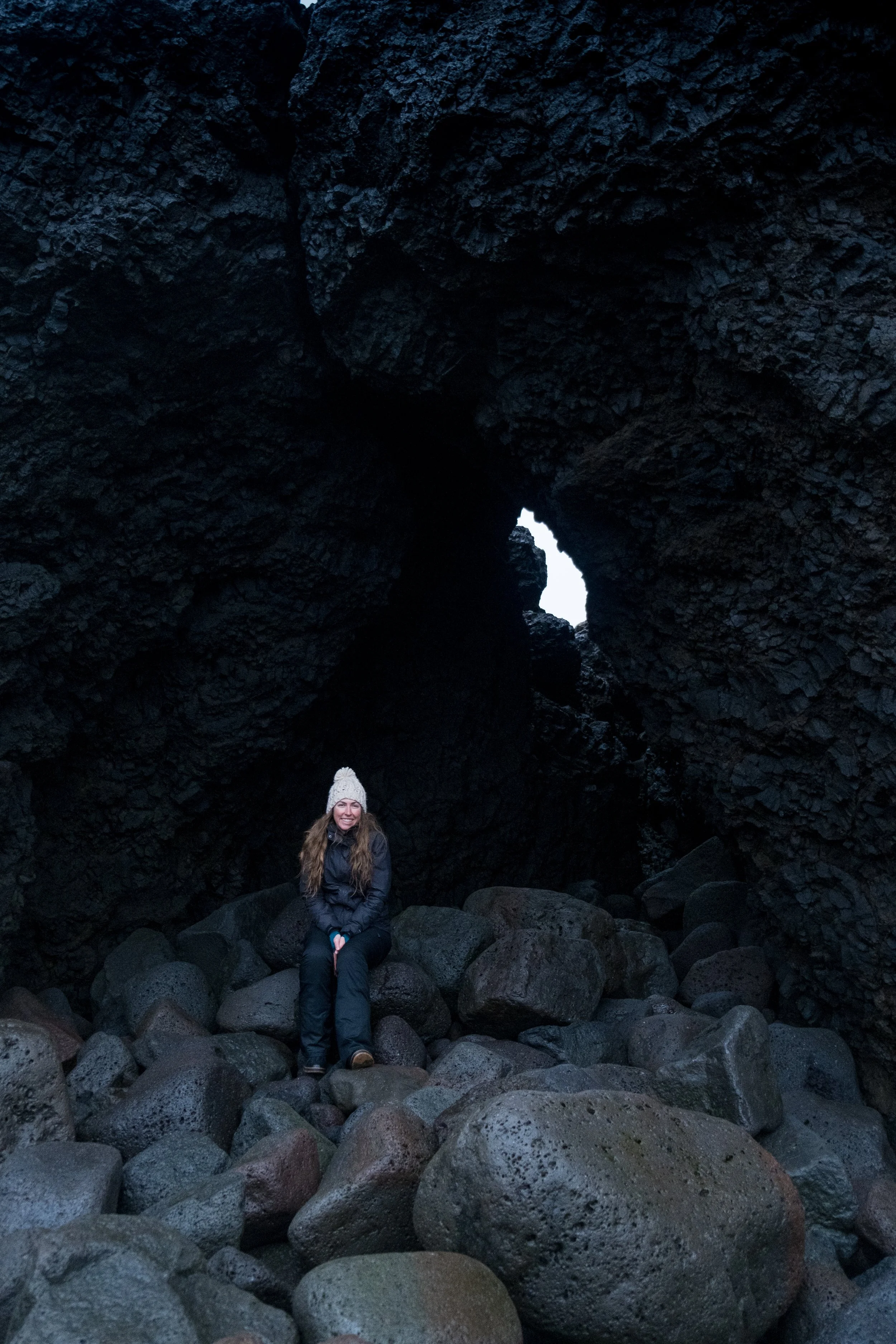 A woman with long hair and a white knit hat sitting on rocks inside a dark volcanic cave with a small opening showing bright sky outside.