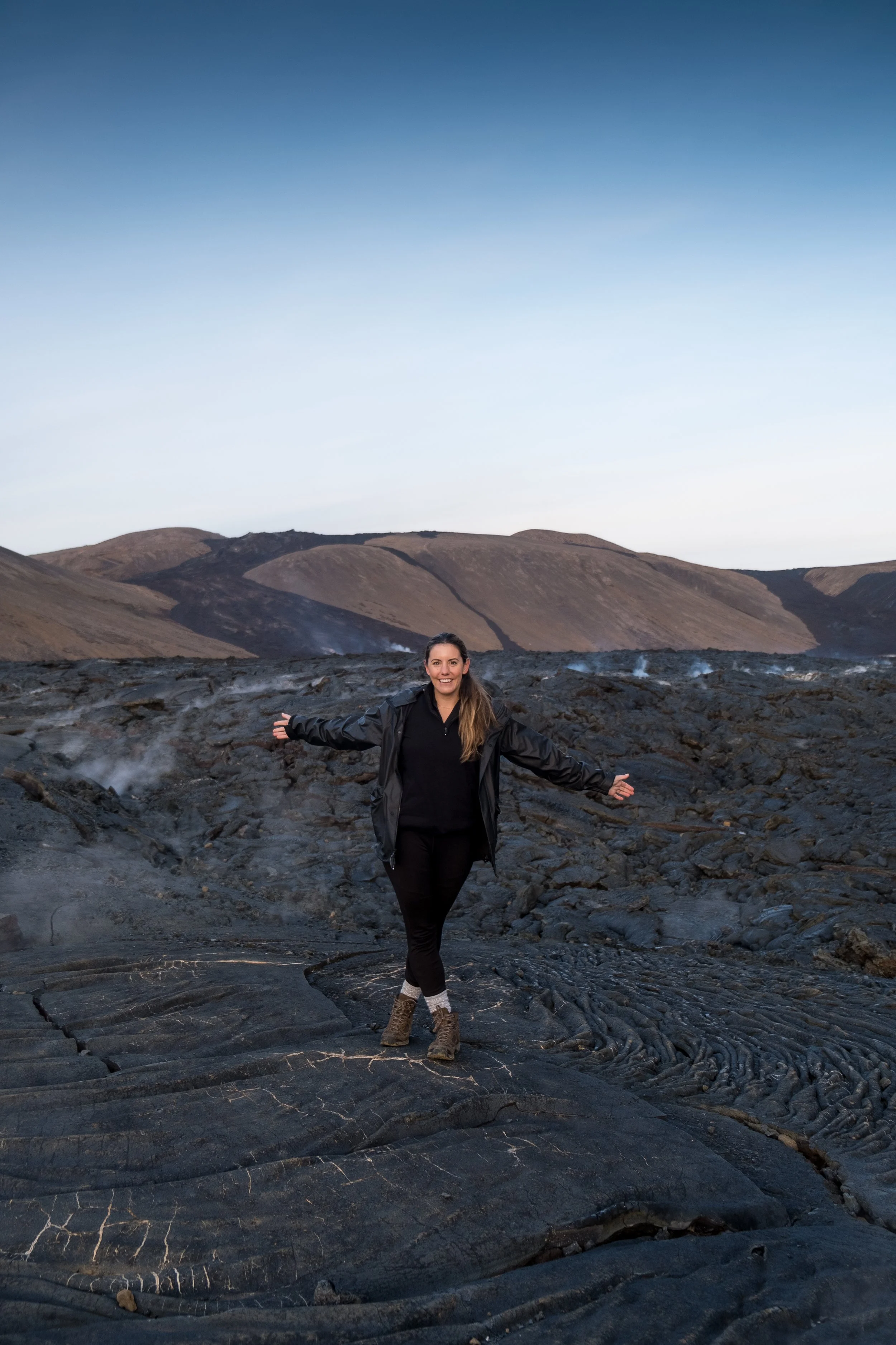 A woman standing on volcanic rocks with her arms outstretched, smiling, mountains in the background, clear sky above.