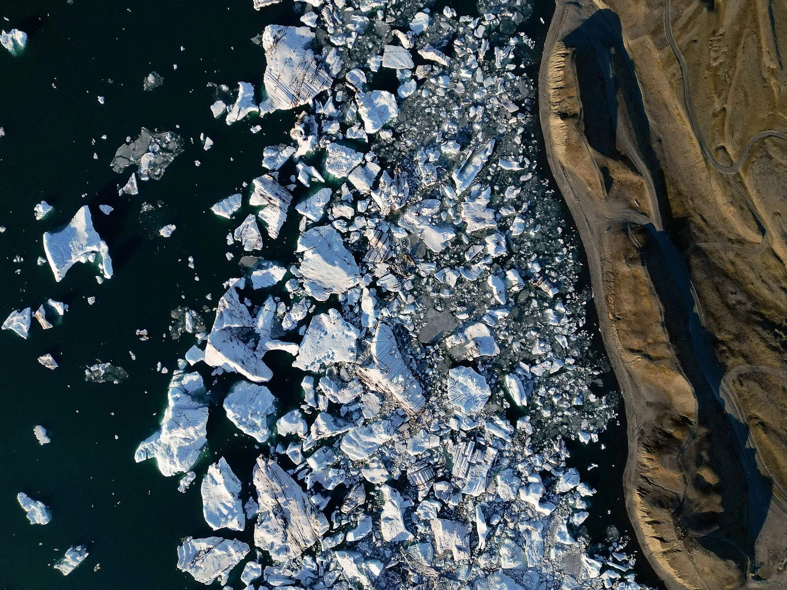 Aerial view of icebergs floating in the ocean near a rocky shoreline and a winding road.