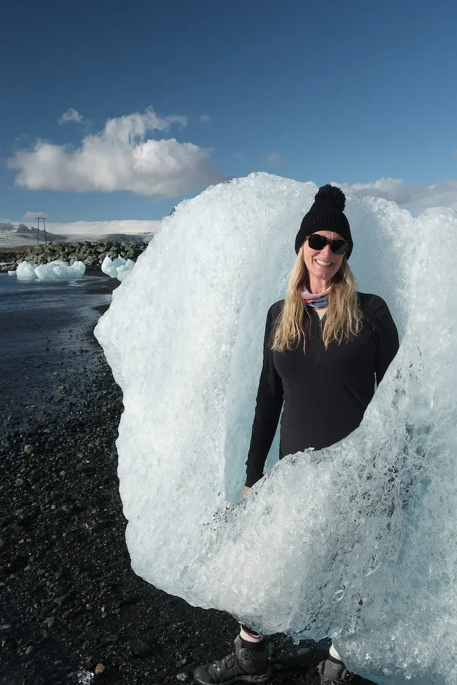 Smiling woman in sunglasses, black hat, and outdoor clothing standing inside a large piece of ice shaped like a frame on a rocky beach with icebergs and cloudy sky in the background.