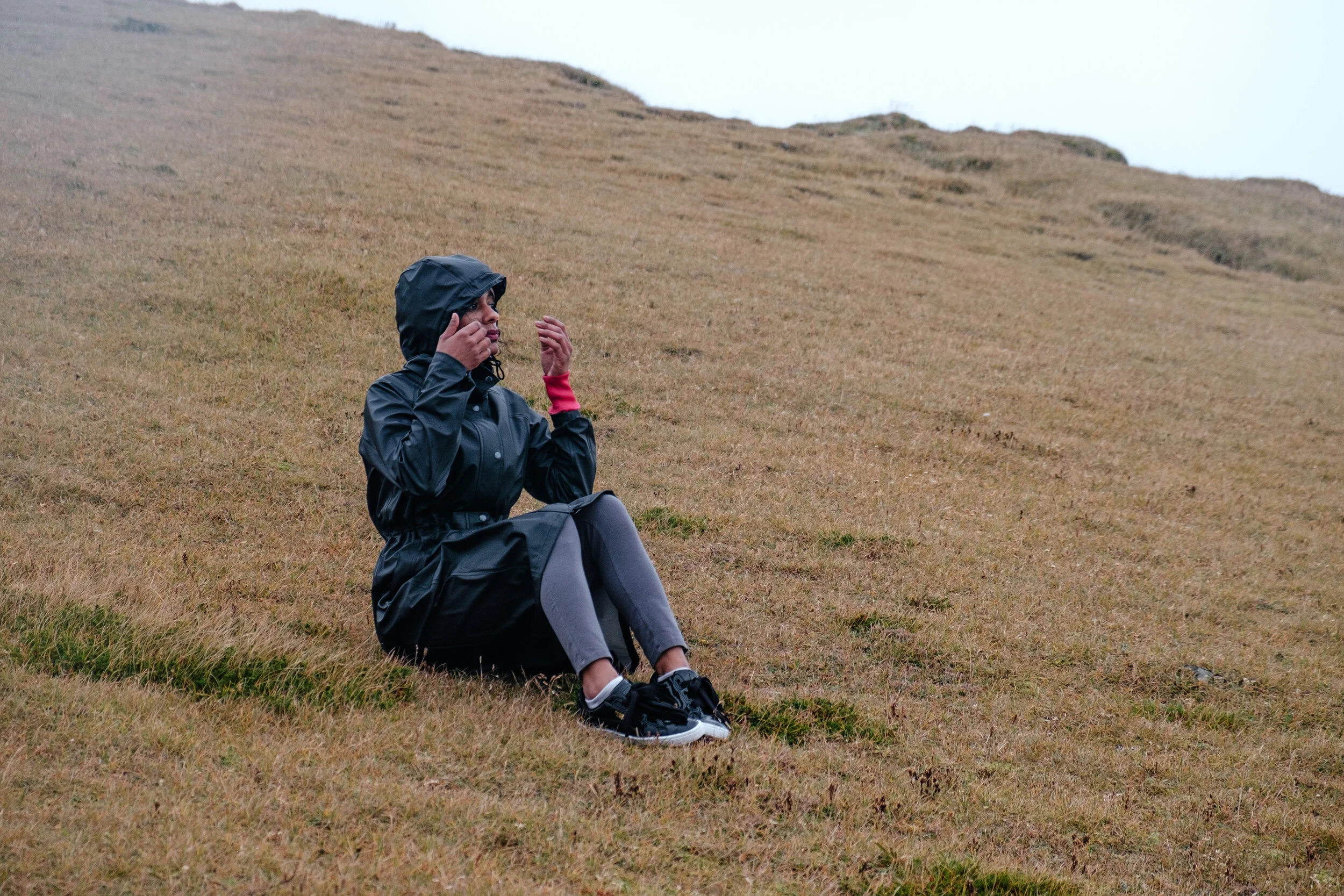 A person sitting on a grassy hillside, wearing a black hooded rain jacket, gray leggings, and black sneakers, with hands adjusting the hood, under an overcast sky.
