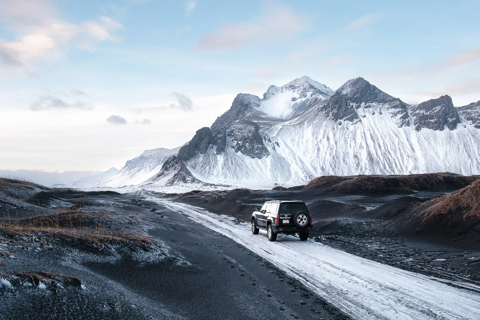 A black SUV driving on a dirt road through a snowy, mountainous landscape with rugged peaks in the background, under a partly cloudy sky.