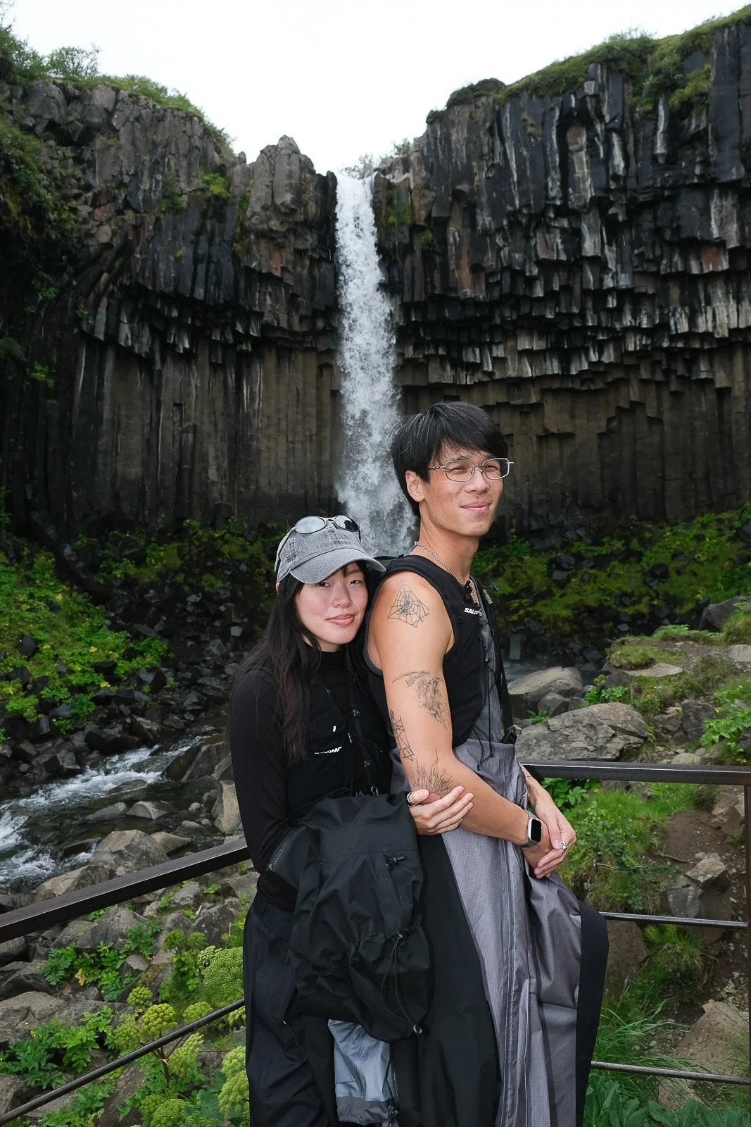 A couple standing in front of a waterfall with basalt columns, surrounded by lush greenery.