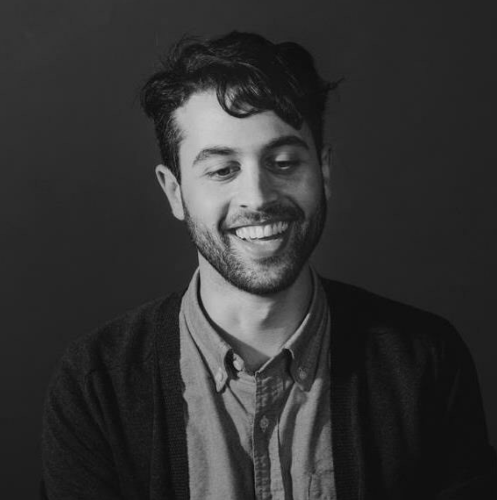 Black and white photo of a smiling man with dark hair, wearing a collared shirt and a cardigan, looking down slightly.
