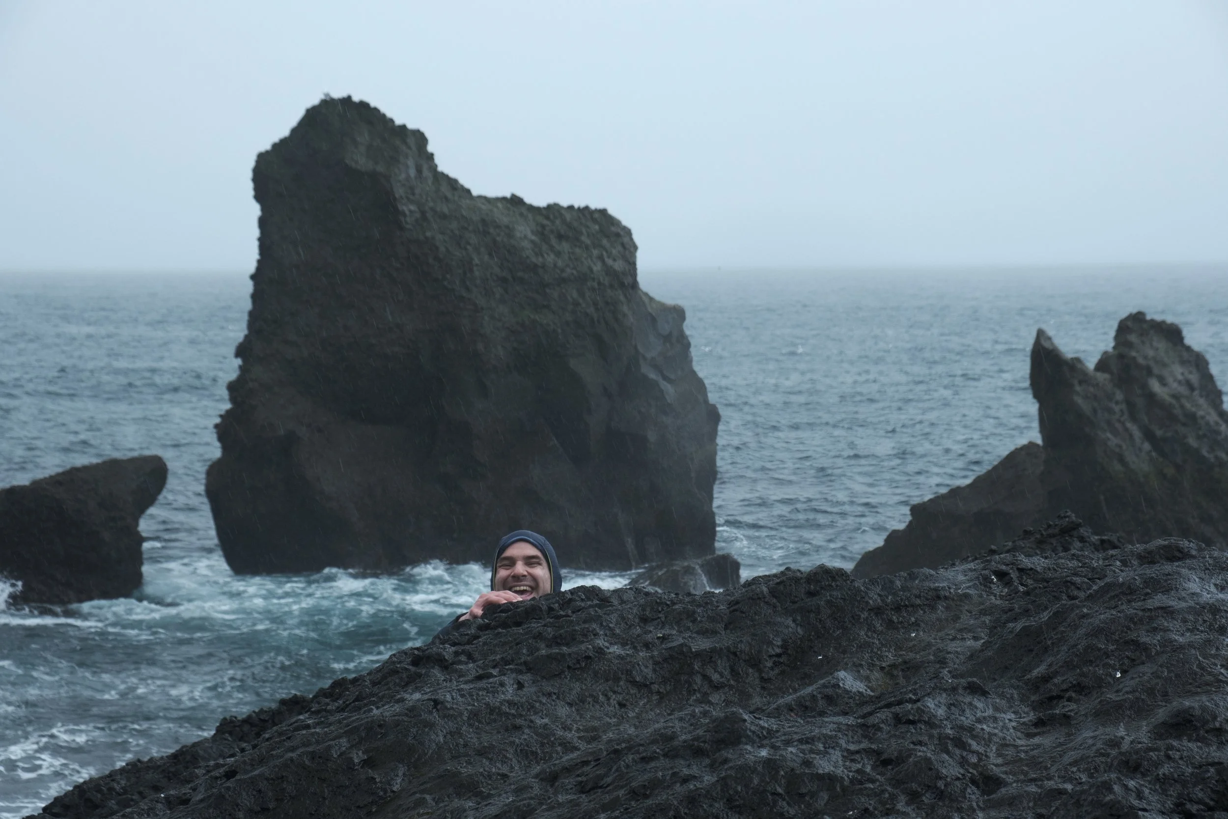 A person smiling and sitting among rocks by the ocean, with large rock formations and a cloudy sky in the background.