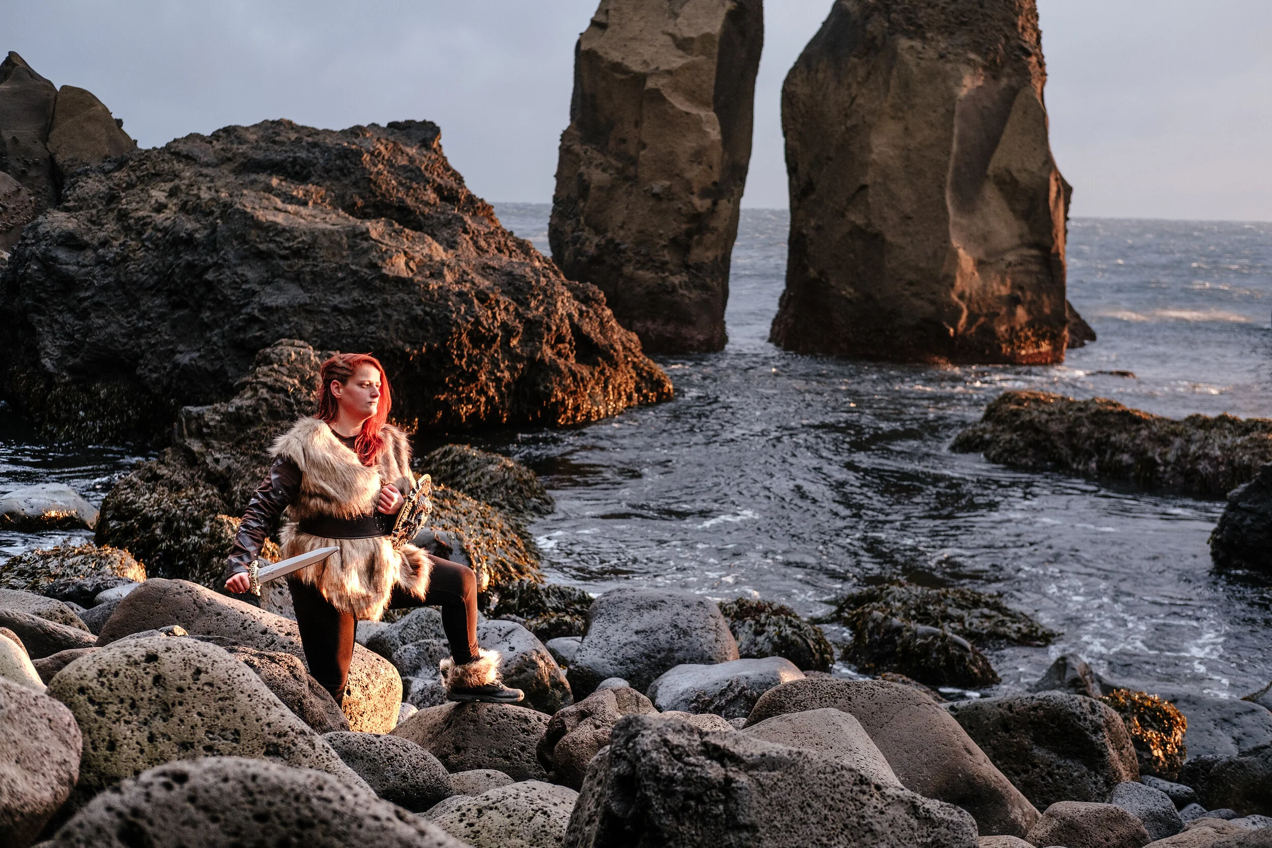 A woman dressed as a Viking warrior, holding a sword, standing on rocks by the ocean with large rock formations in the background during sunset.
