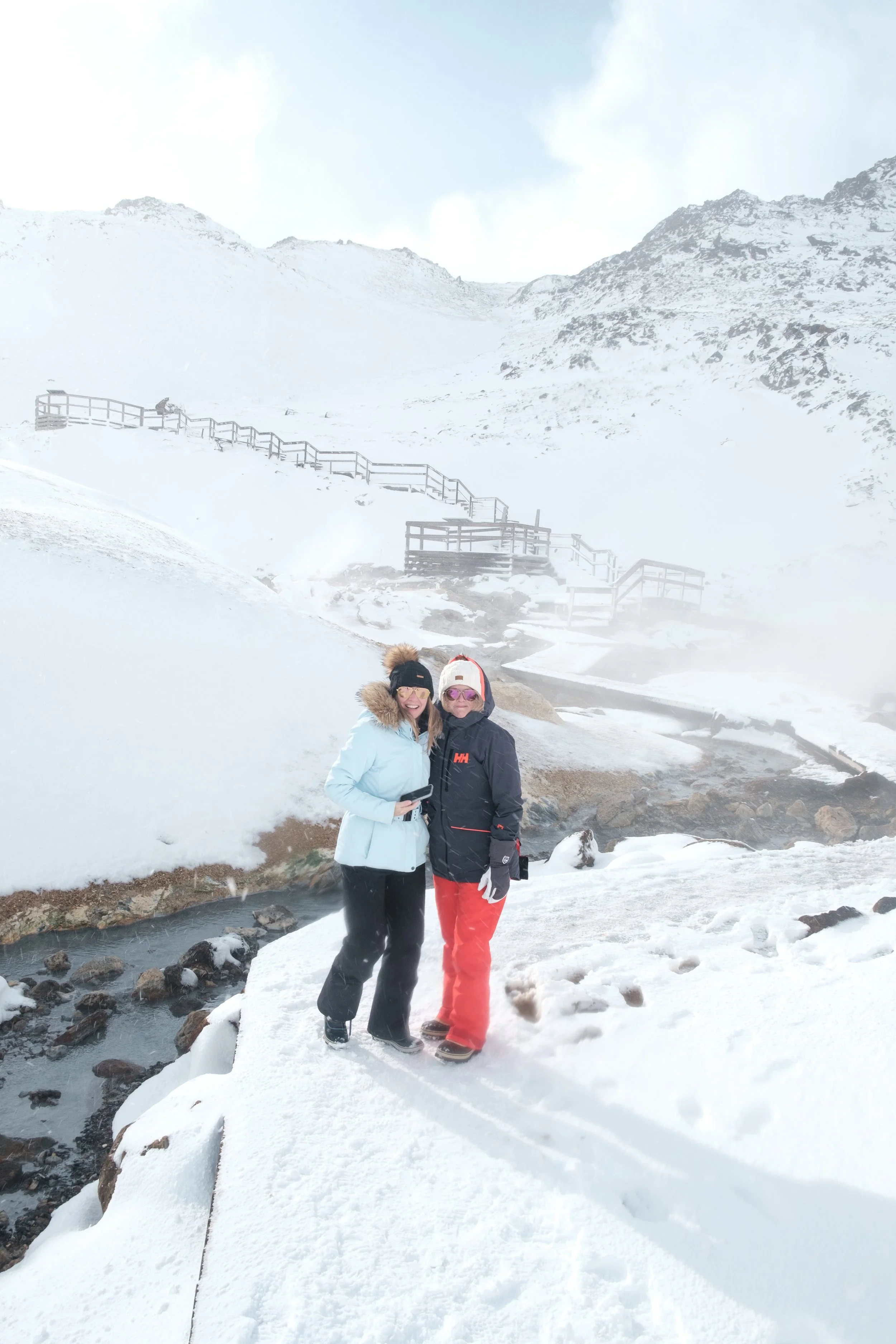 Two women in winter clothing standing on a snow-covered landscape with mountains in the background, near a small stream.
