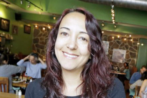 A woman with wavy, shoulder-length hair smiling in a restaurant with green walls, stone accents, and string lights, while other people dine in the background.