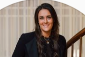 A woman with long dark hair, wearing a black blazer and patterned blouse, smiling in front of a staircase.