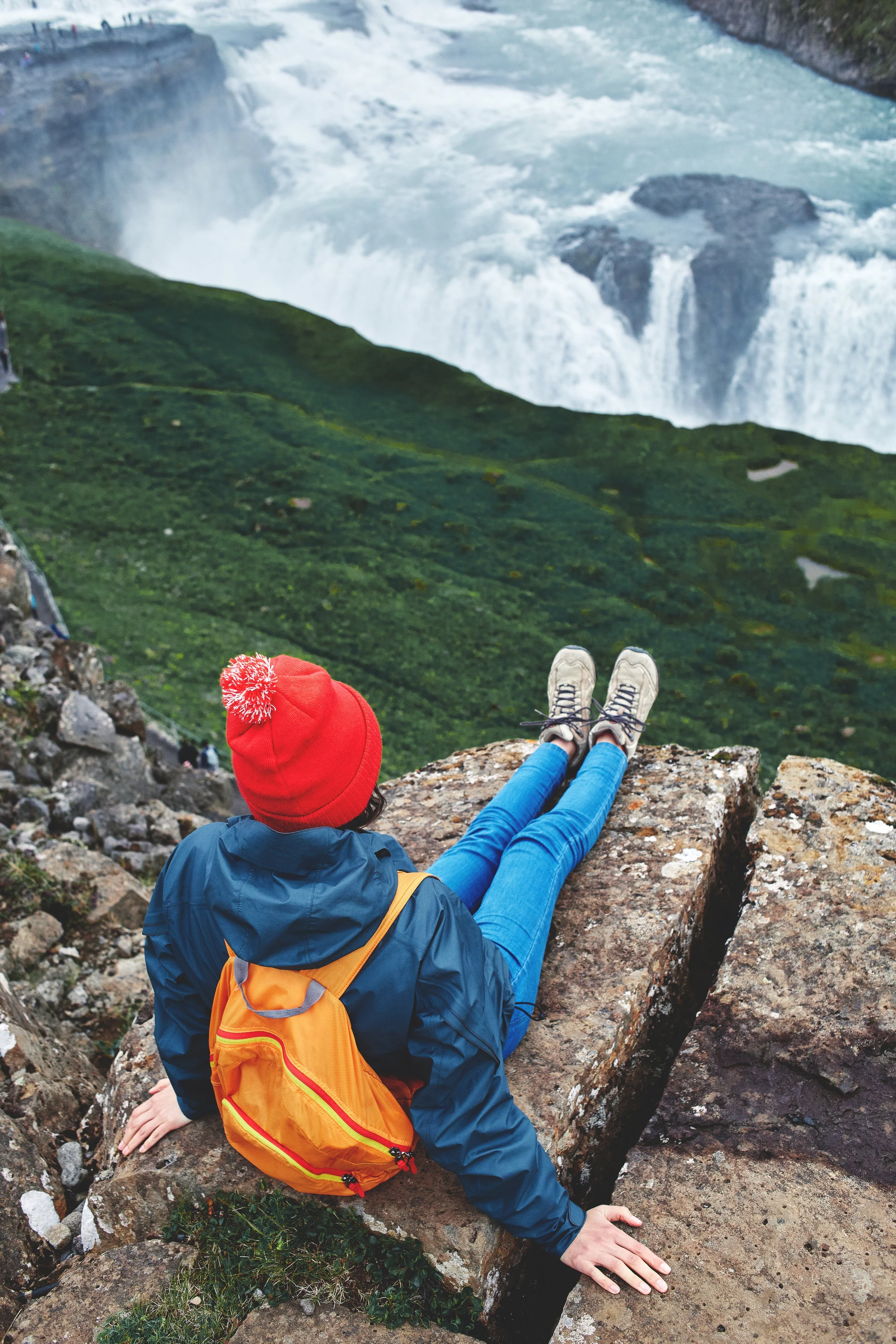 Person in a red hat and blue jacket sitting on a rocky ledge, on Gullfoss Waterfall