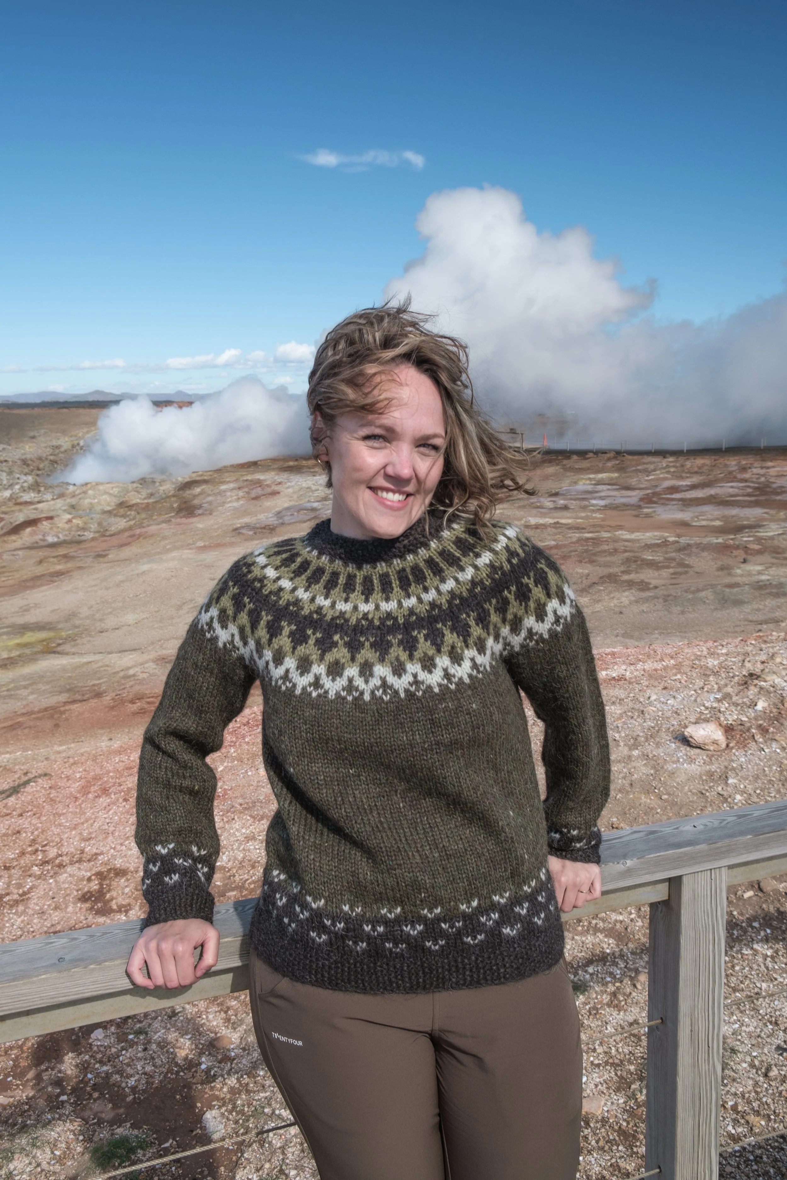 A woman with wavy blond hair smiling outdoors, wearing a patterned sweater and brown pants, standing near a wooden railing with geothermal steam and a blue sky in the background.