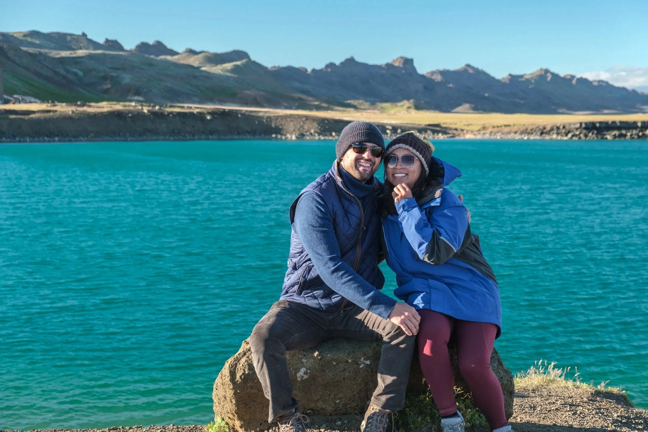 A couple sitting on a large rock by a turquoise lake, surrounded by mountains, wearing sunglasses and warm clothing, smiling at the camera.