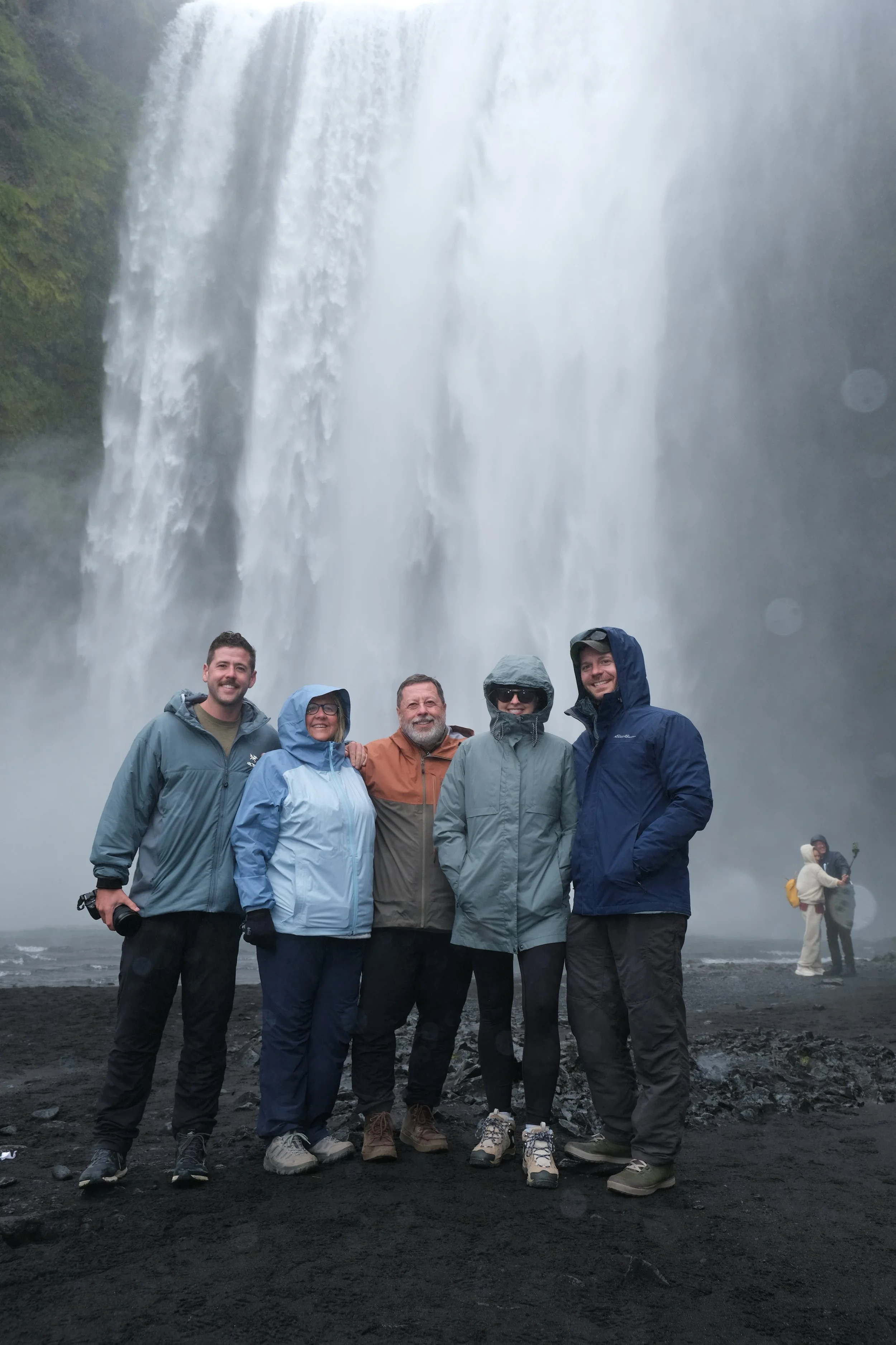 A group of six people standing in front of a large waterfall, all wearing rain jackets and outdoor gear, with two other people in the background taking photos.