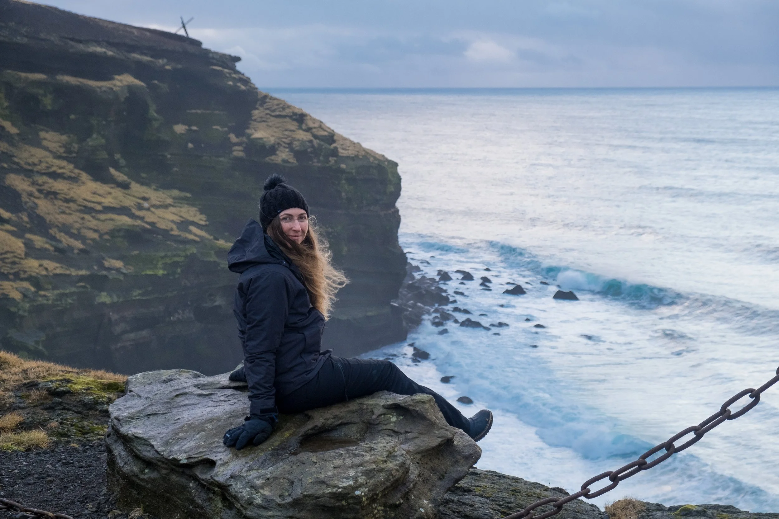 A woman dressed in a black jacket and a knit hat with a pom-pom, sitting on a large rock near the ocean at a cliffside. She is smiling and looking at the camera with dark hair blowing in the wind. The background features a grassy cliff, rocky shoreli